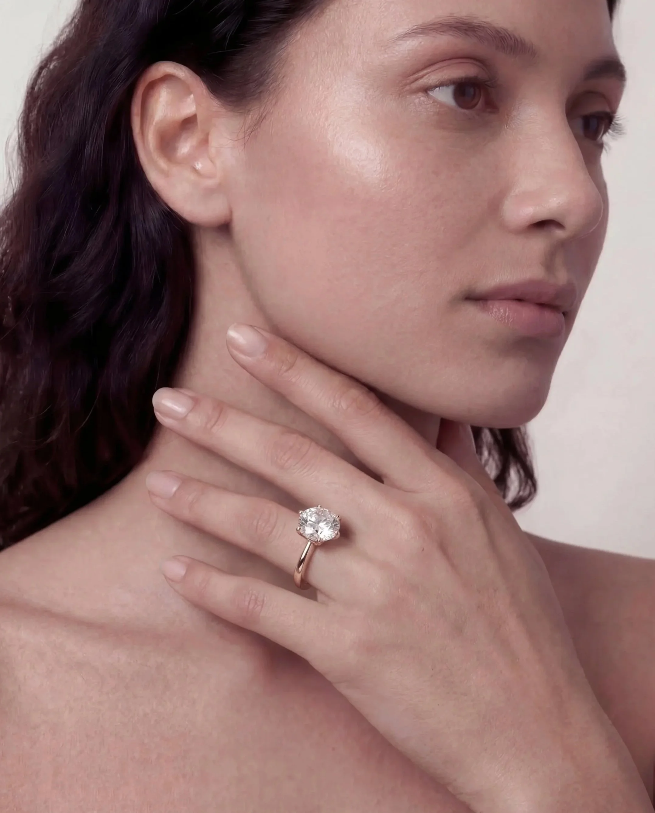 A woman with fair skin and dark brown hair, wearing a large diamond ring on her left hand, gently touching her neck wearing a diamond ring.