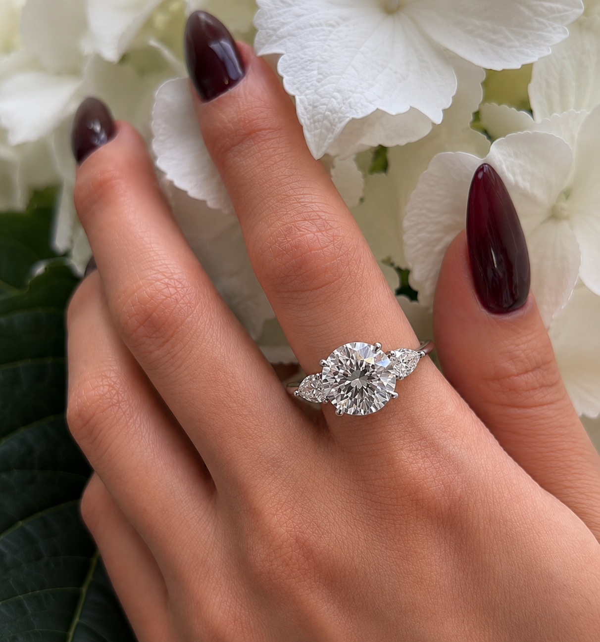 A hand with dark red nails wearing a diamond engagement ring with two pear-shaped side stones, set against white flowers and green leaves.