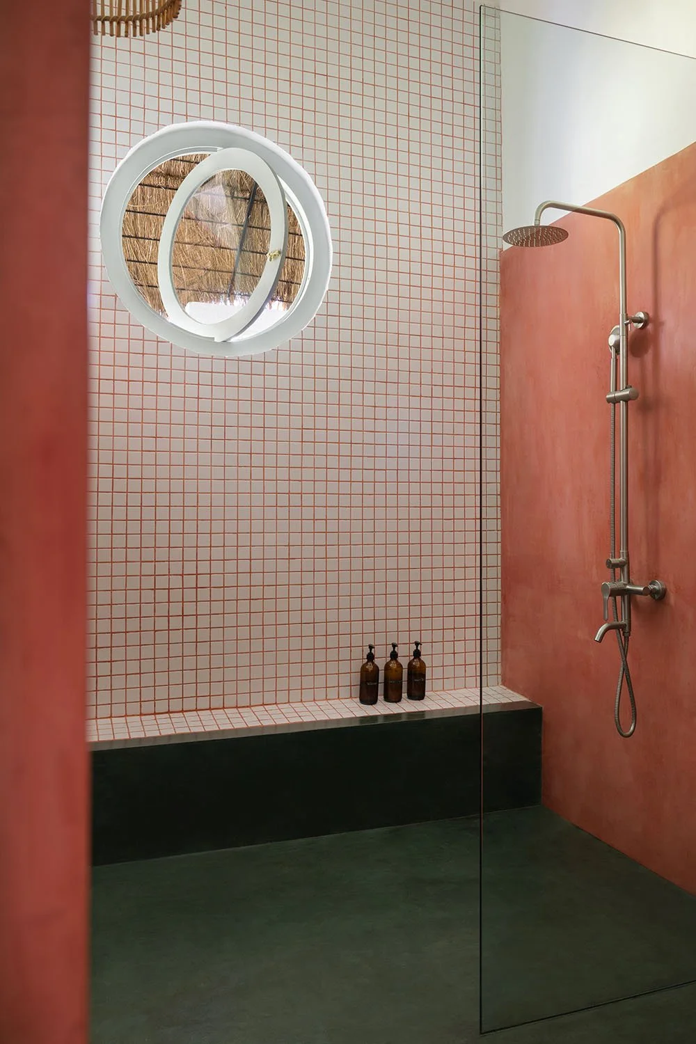 Minimalist walk-in shower with rised pink wall, white and orange grid-tiled wall, black base, and small bottles on bench, reflecting in mirror.