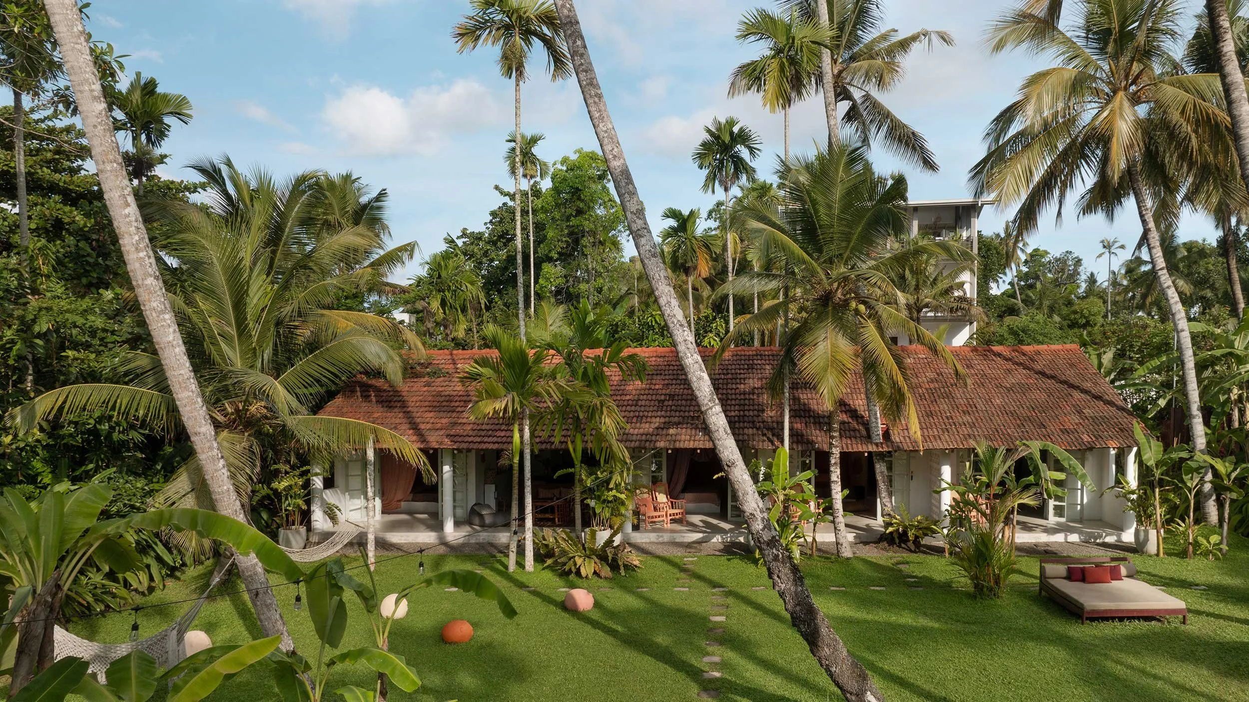 A thatched roof house surrounded by lush greenery and tall palm trees, with a patio area and outdoor seating, under a partly cloudy sky in a tropical setting.
