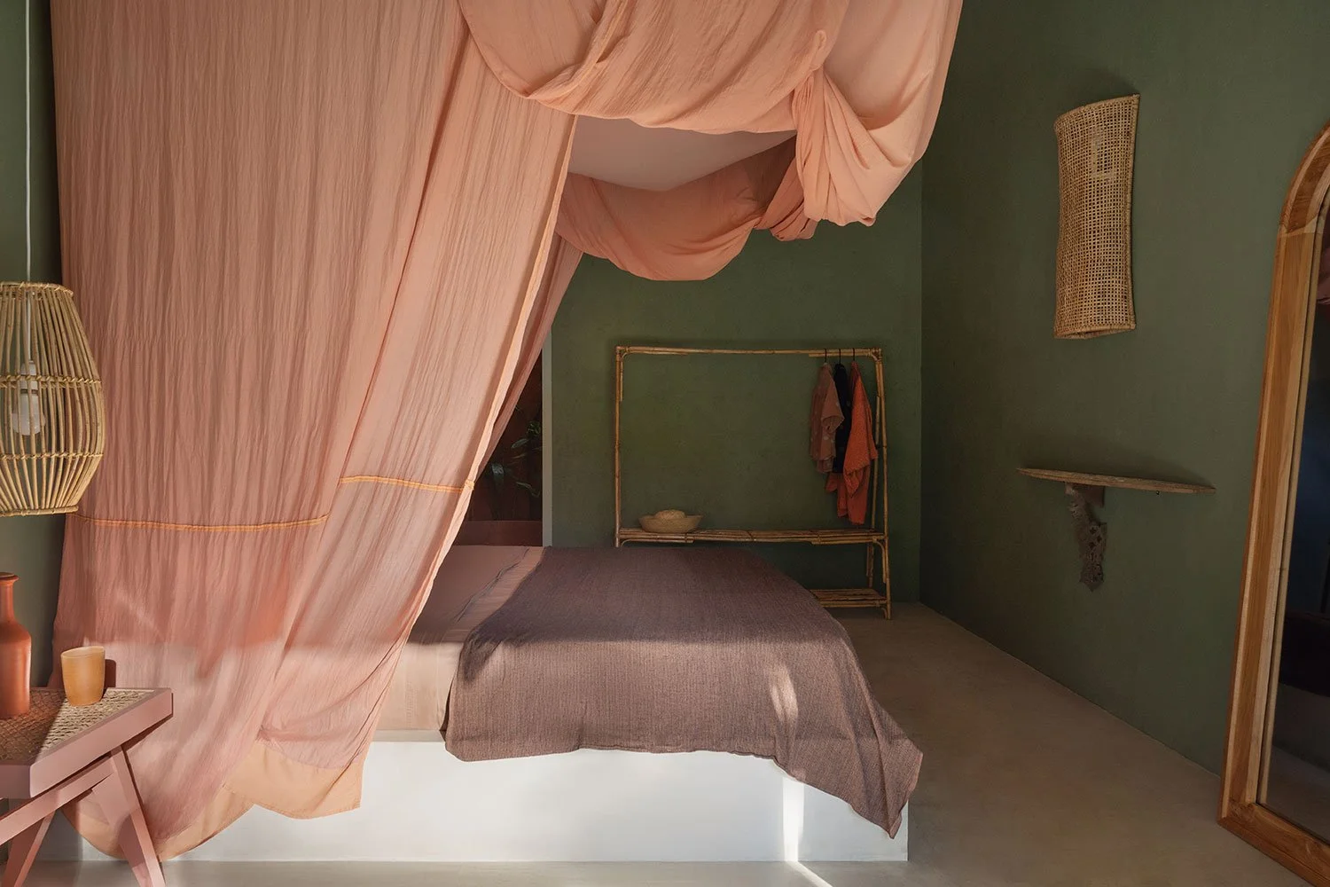 A bedroom with a bed draped in pink canopy and brown bedding. Green walls with rattan wall decor and shelf, bamboo frame with hanging clothing. Soft lighting and earthy decorations.
