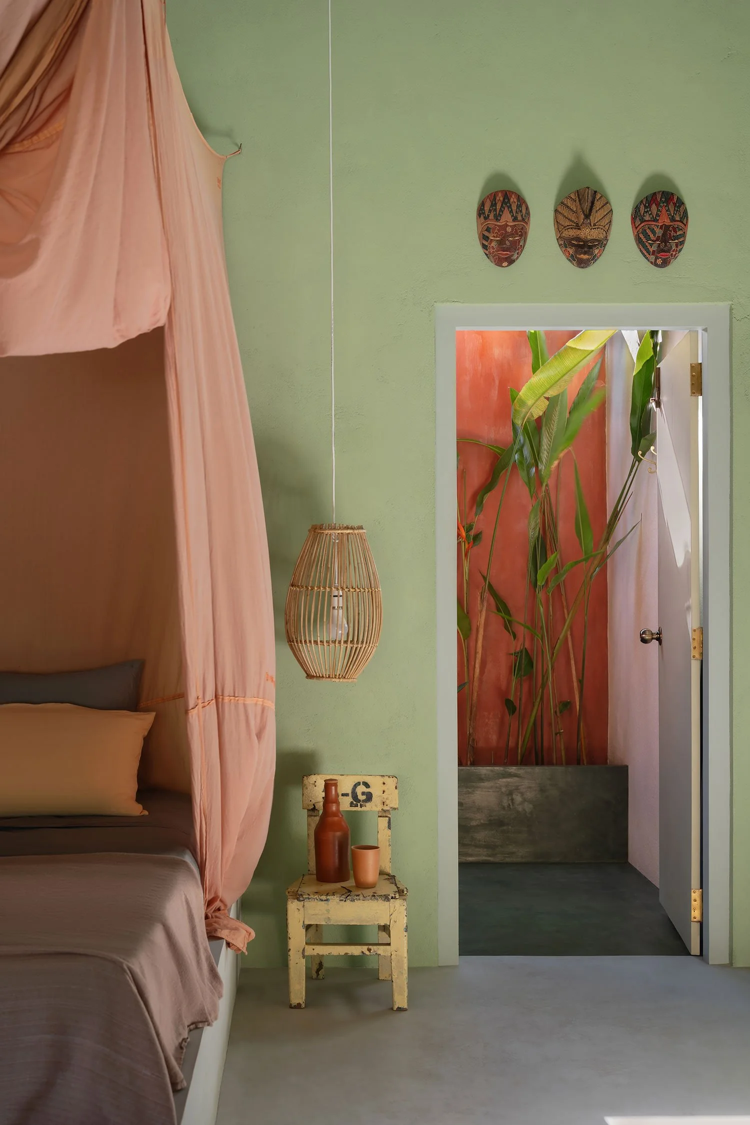 Interior of a bedroom with a bed on the left, separated by pink curtains. The wall is painted light green and decorated with three colorful tribal masks. A small distressed white table holds a brown bottle and a cup. There's a doorway revealing a room with tall green plants and stands out with a reddish wall.
