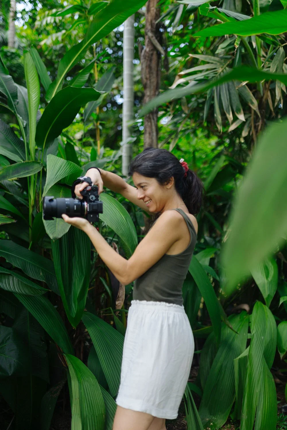 A woman with dark hair tied back, wearing a dark green tank top and white shorts, is smiling and taking photos with a professional camera while surrounded by lush green tropical plants — Casa Tikiri, Ahangama, Sri Lanka
