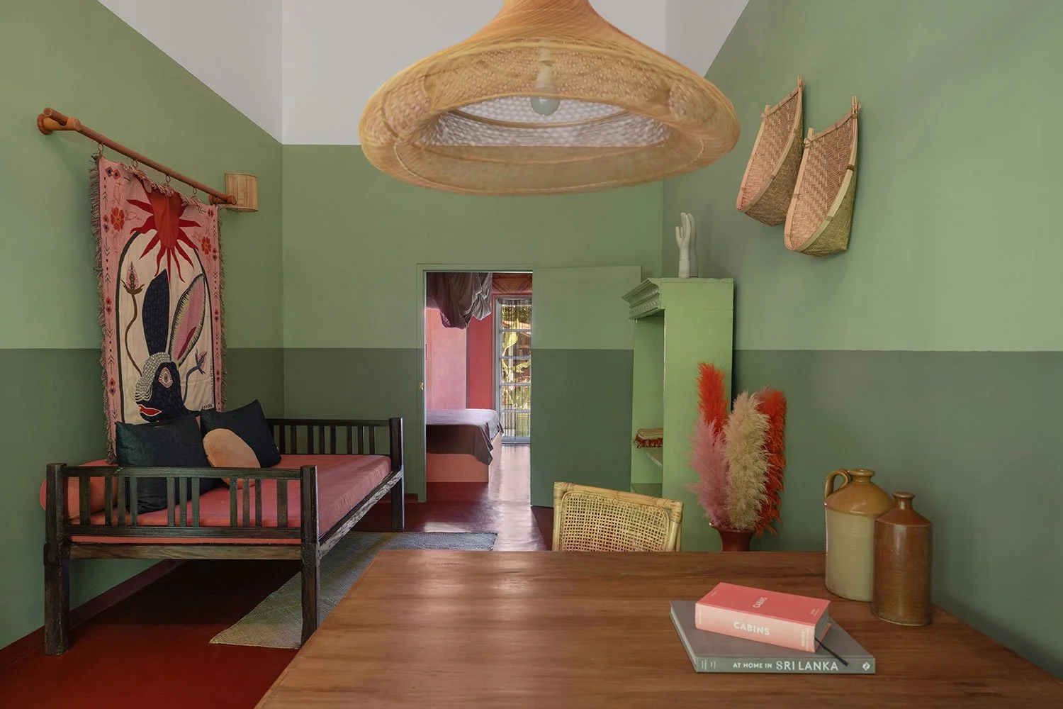 A cozy room with green walls, a wooden table with books and vases, a daybed with pillows, hanging baskets, a woven lamp, and a doorway leading to a bedroom with a balcony.