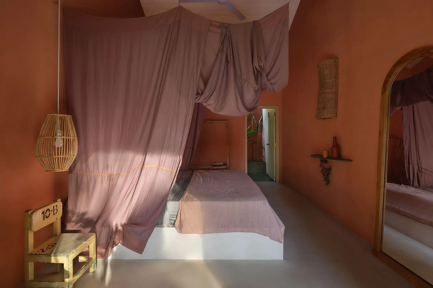 A bedroom with pink walls, featuring a bed with pink bedding and pink curtains hanging above it. There is a small wooden chair with yellow paint and markings, a rattan pendant light, a mirror on the right wall, a small shelf with decorative items, and an open door leading to a hallway with plants.