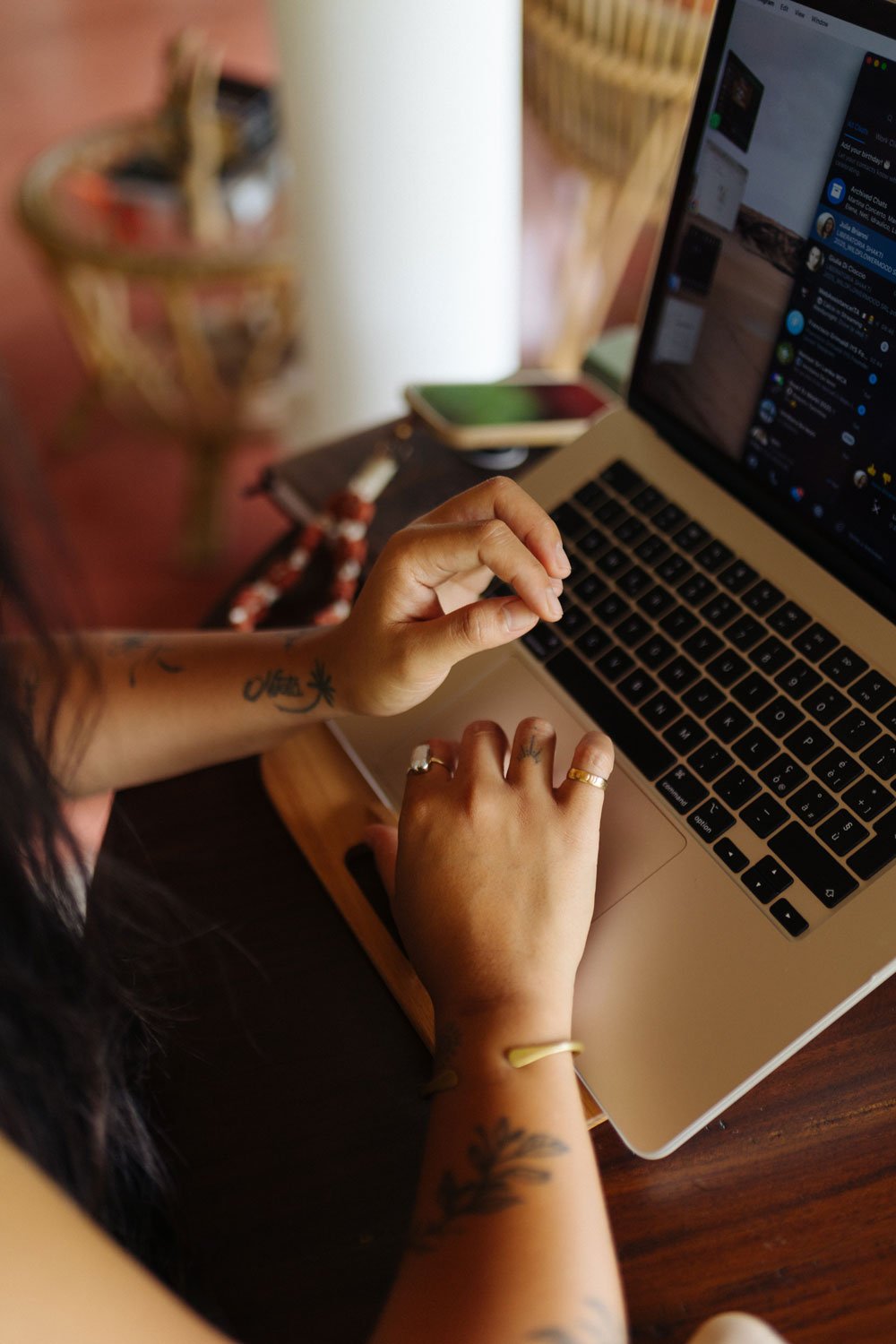 Person using a MacBook computer at a wooden table, with a smartphone and a notebook nearby, in a cozy indoor setting — Casa Tikiri, Ahangama, Sri Lanka