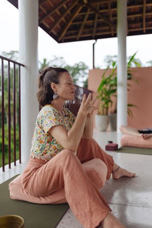A woman practicing yoga outdoors on a patio, sitting cross-legged on a yoga mat with her hands in a prayer position, surrounded by plants and a wooden roof — Casa Tikiri, Ahangama, Sri Lanka