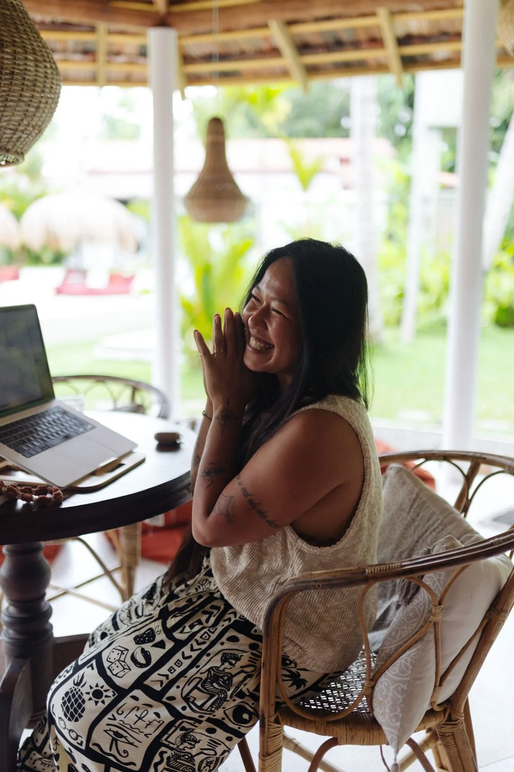Woman sitting at a wooden table with a laptop, outdoors in a tropical setting, laughing with her hand near her face — Casa Tikiri, Ahangama, Sri Lanka
