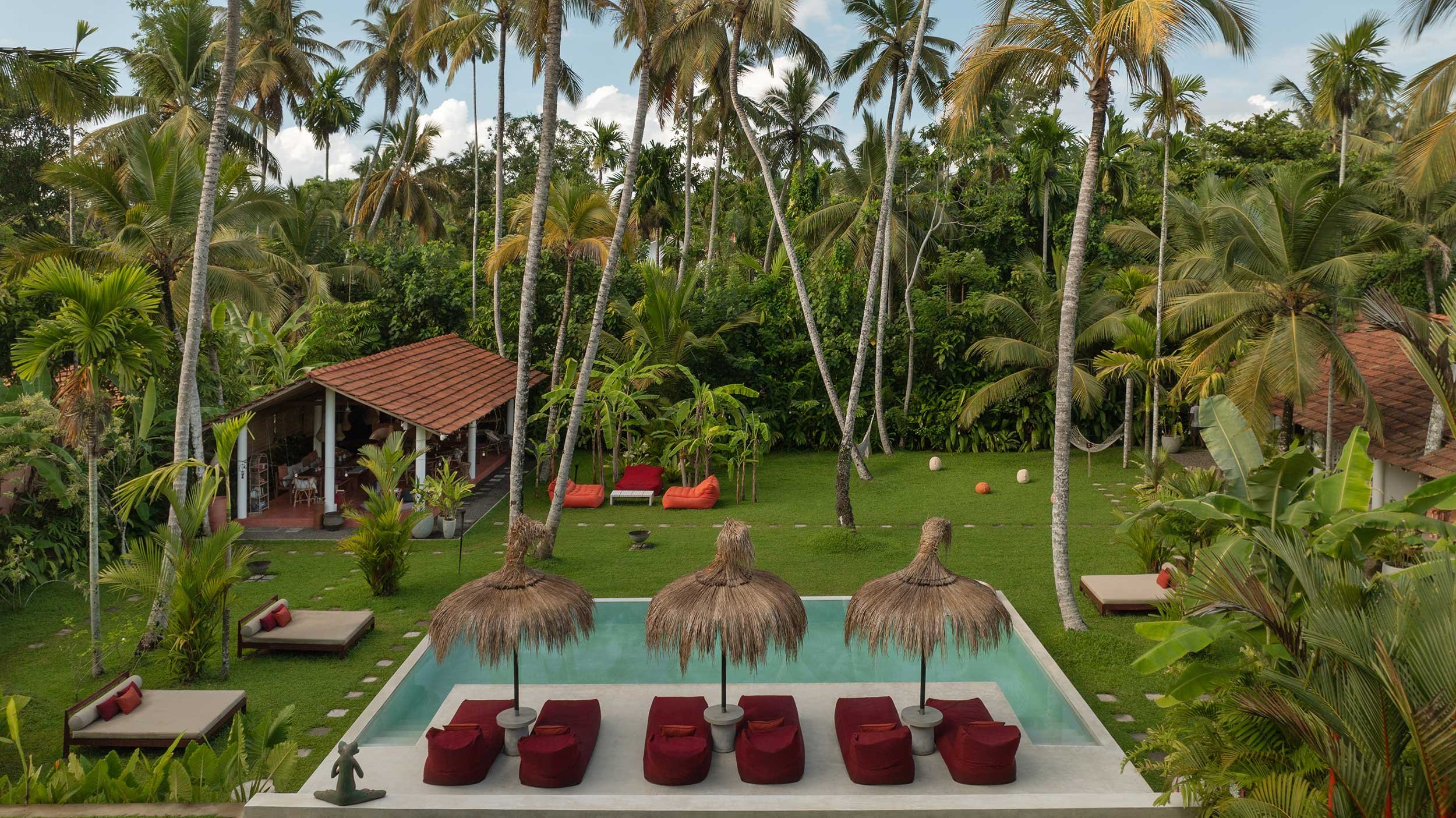 Tropical resort with a swimming pool shaded by thatched umbrellas, surrounded by green lawn, tall palm trees, and a small open-air structure with red-tiled roof, outdoor seating, and lounge chairs with cushions.