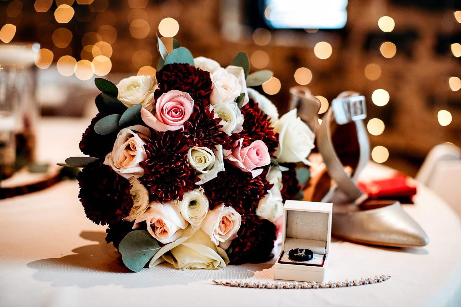 A bridal bouquet with pink, white, and dark red roses and greenery, a diamond ring in a white box, a pearl necklace, a vintage high-heeled shoe, and a red cloth, all arranged on a table with a blurred background of warm lights.
