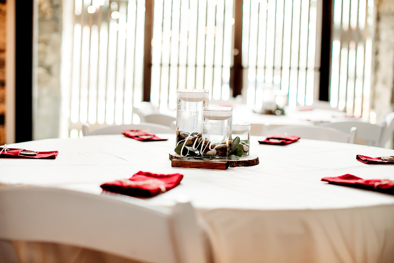 Table set for an event with a white tablecloth, around it are white chairs, and red napkins with silverware. The centerpiece features three clear cylindrical vases filled with water, rocks, and greenery, with a wooden base and a sign that says "Love"