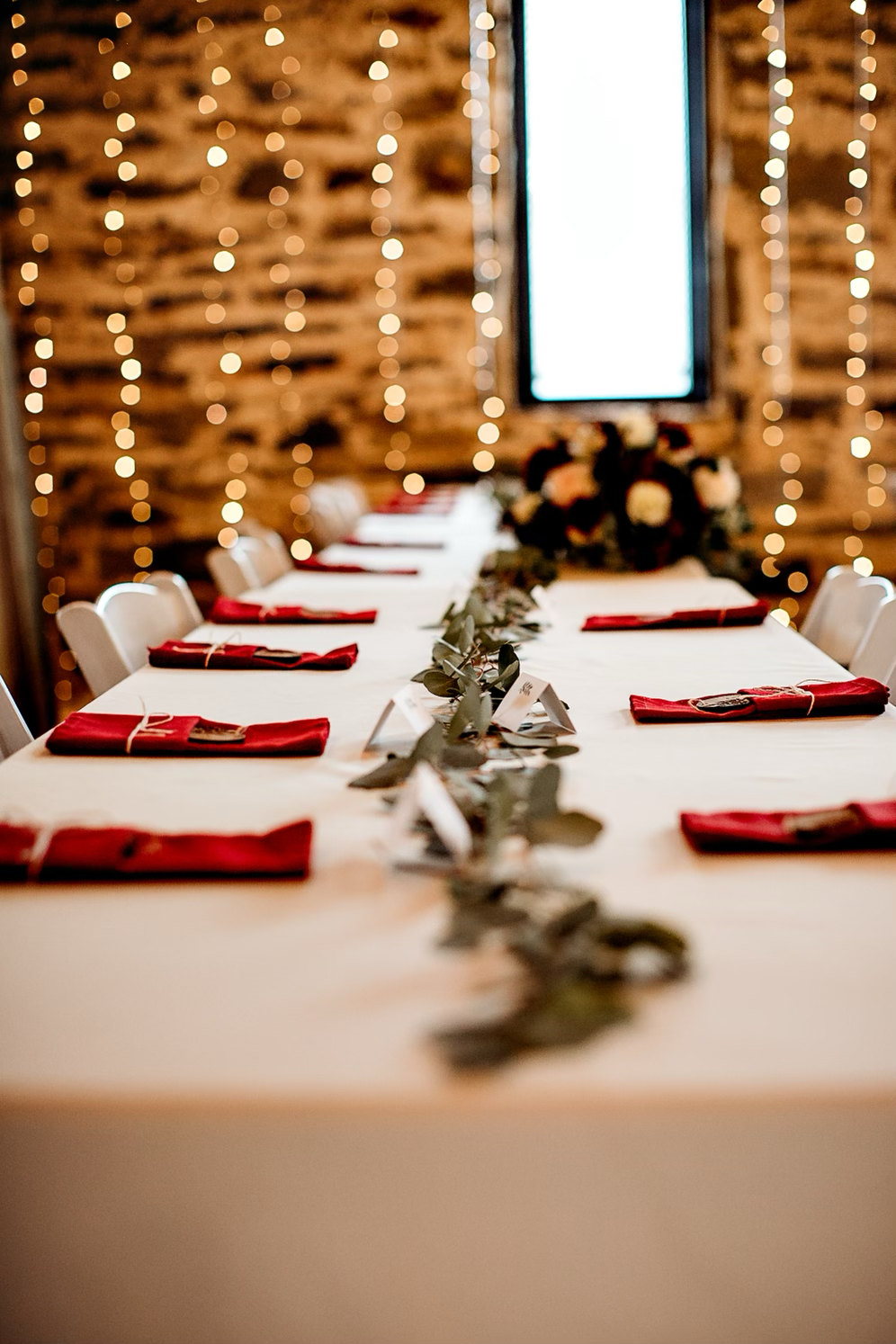 A long rectangular dining table set for an event with red napkins, a leafy table runner, and a floral centerpiece, against a rustic wooden wall with string lights and a window in the background.