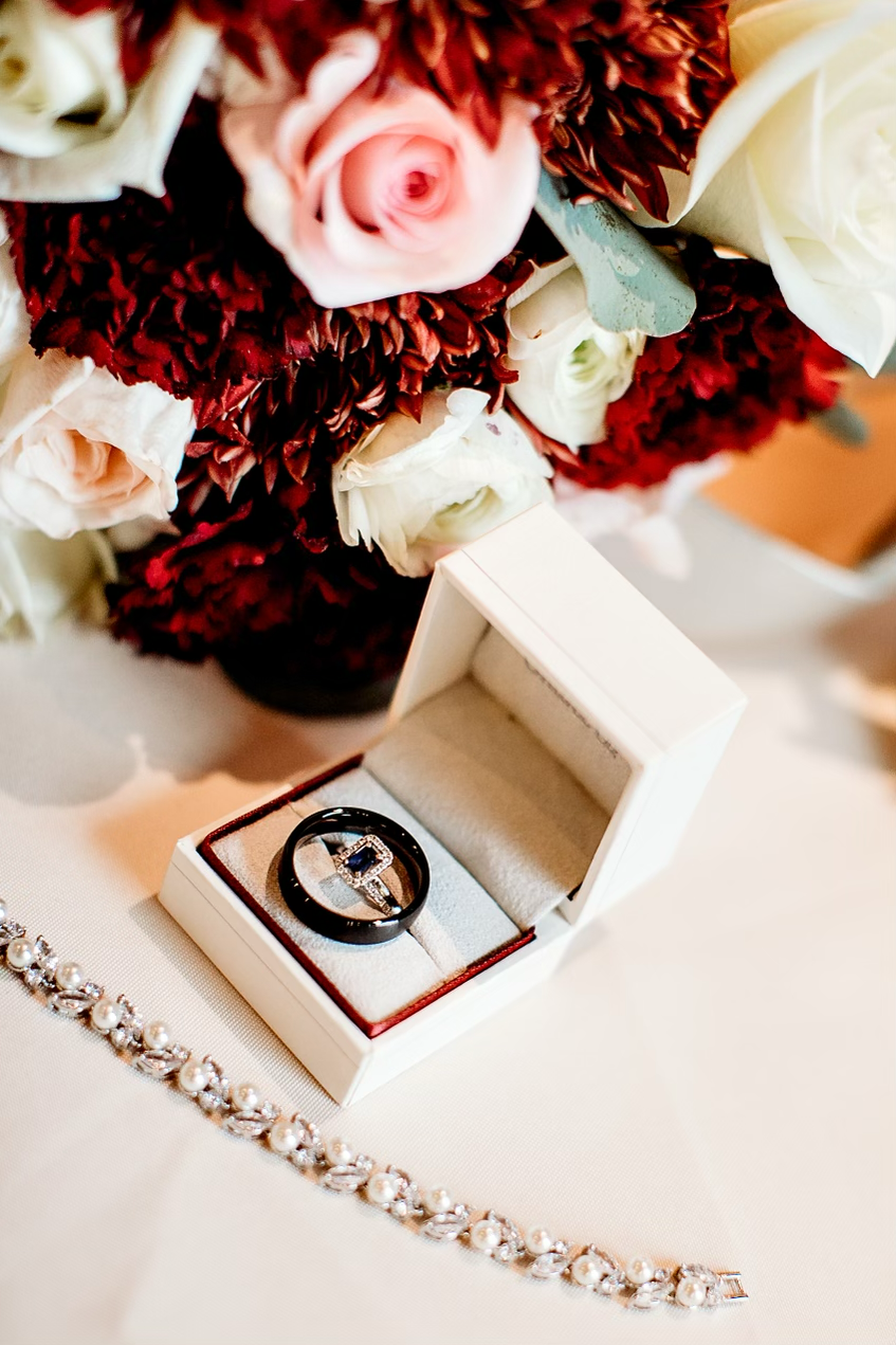 A jewelry box with a ring inside, a pearl bracelet, and a bouquet of flowers including pink and white roses and burgundy flowers.