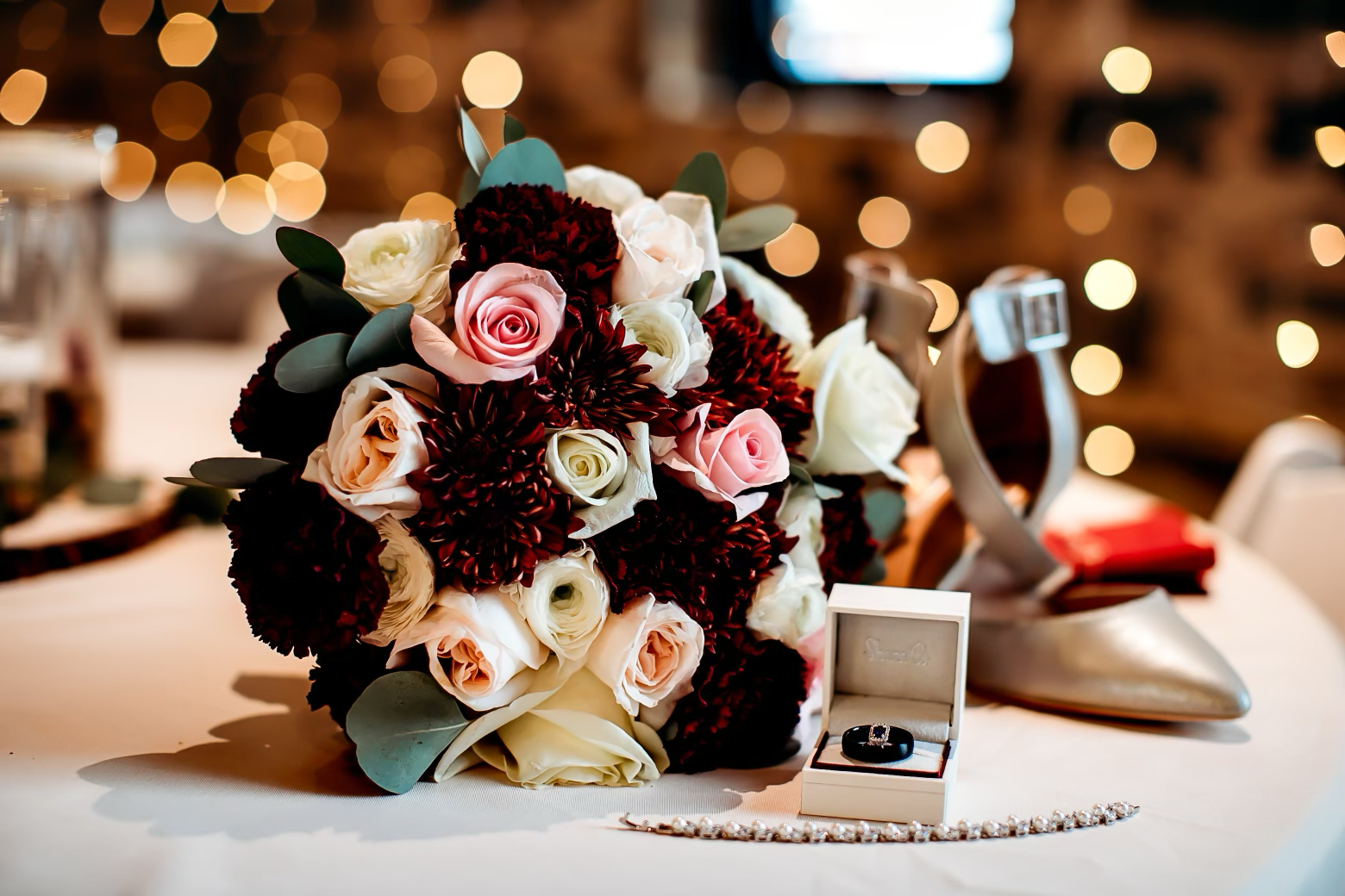 A wedding bouquet of pink, white, and burgundy flowers, a black engagement ring in a white box, a pearl bracelet, and a gold-colored shoe with a red cloth inside, all on a white table with blurred warm lighting in the background.