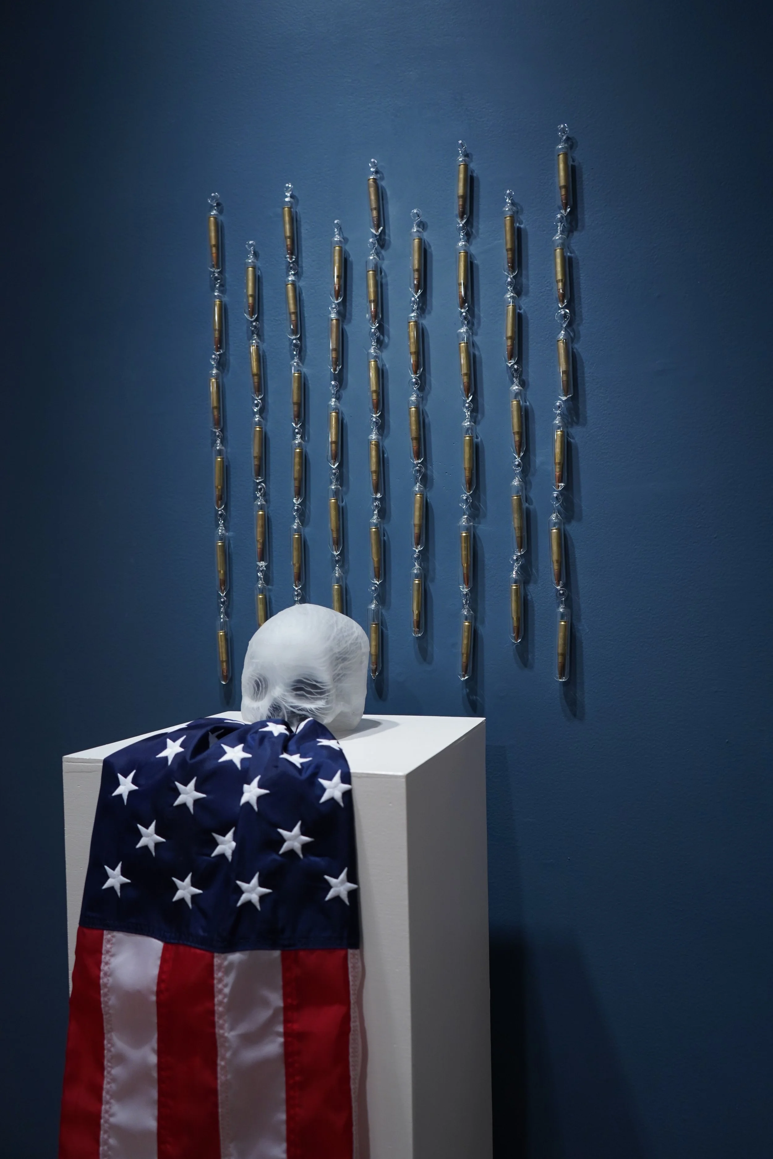 ...Wrapped in a Flag and Bearing Munitions. 2026. Hot blow molded glass, American flag, flameworked glass, M60 ammunition