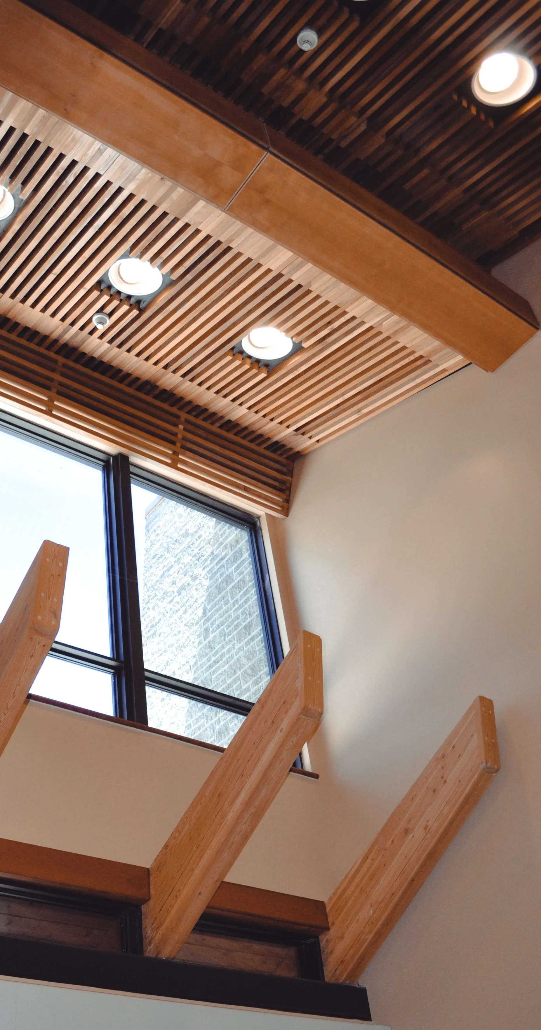 Custom wooden accents on the ceiling of a hospital with thin wooden panels going vertically leading into horizontal panels above large windows. Pergola shaped beams protrude from the wall below the windows to create depth
