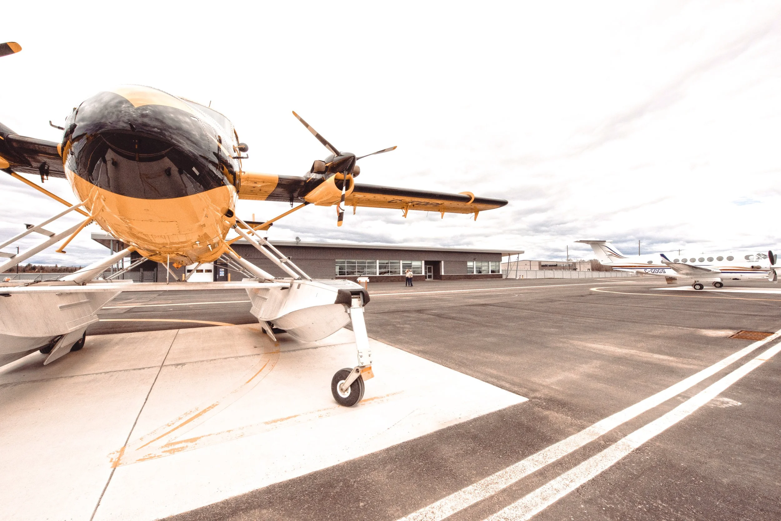 Yellow Airplane sitting on landing pad surrounded by large cement airport landing strip and modern, flat, grey building behind.