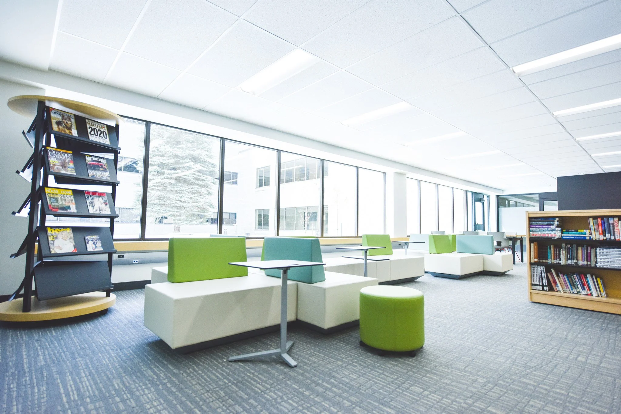 Modern, bright learning commons with tall windows bringing natural light in. White benches with a mix of pale blue and bright green backs. Minimal grey moveable single desks located throughout commons and two wooden bookshelves full of books.
