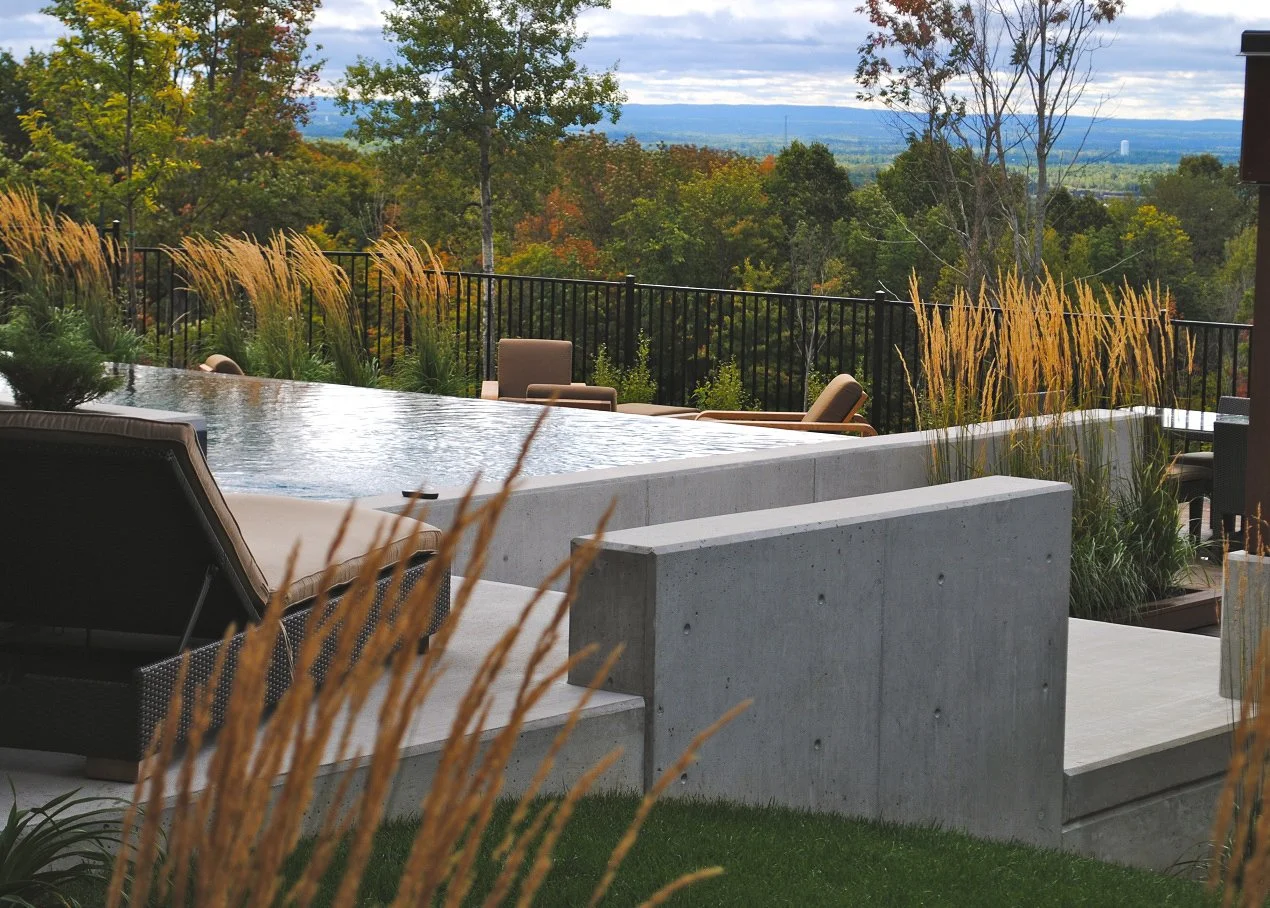 Modern cement slabs in a residential landscaping project surrounding an in the ground pool overlooking forest with goldenrod plants around the pool next to pool chairs