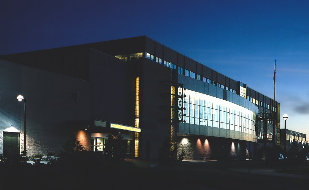 Campus at dusk with lights shining through the long line of windows in the middle of the building with a line of rectangular windows lining the top of the building