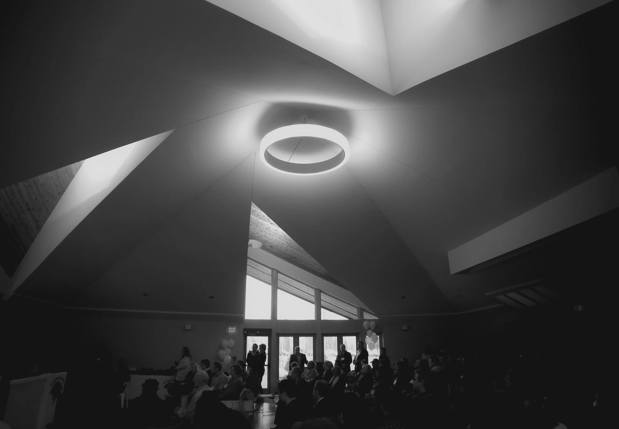 Black and white photo of the inside of a convention centre with people seated in rows, tall abstract ceilings, and a modern, round hanging light in the center of the room