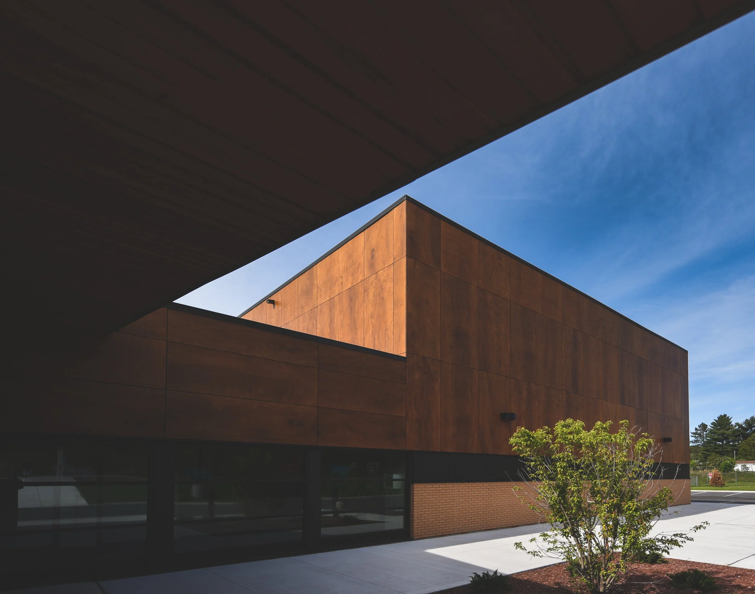 Catholic School with custom wooden siding around the top 75% of the boxed shaped building and dark floor to ceiling windows along the bottom 25% of the building with clean cement walkways and mulch landscaping in the center of the pathways