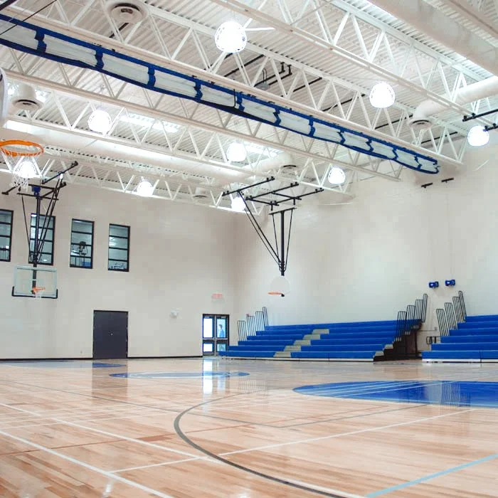 Tall ceilings with white vents and metal framework, two basketball hoops hang from the ceiling, blue bleacher seats on the far right hand side, custom wood court floor with painted instruction lines