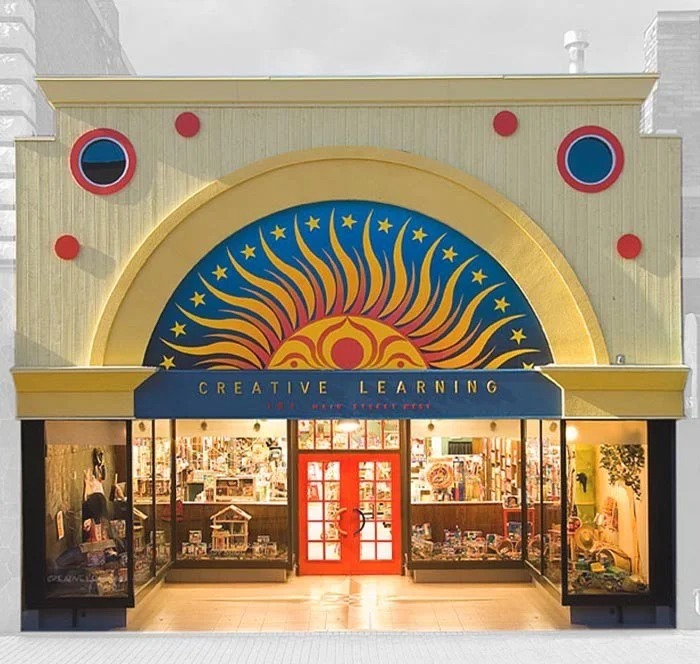 Creative learning toy store with yellow framework around sun logo painted on blue background. Red doors in the middle of floor to ceiling windows with toy displays in the windows