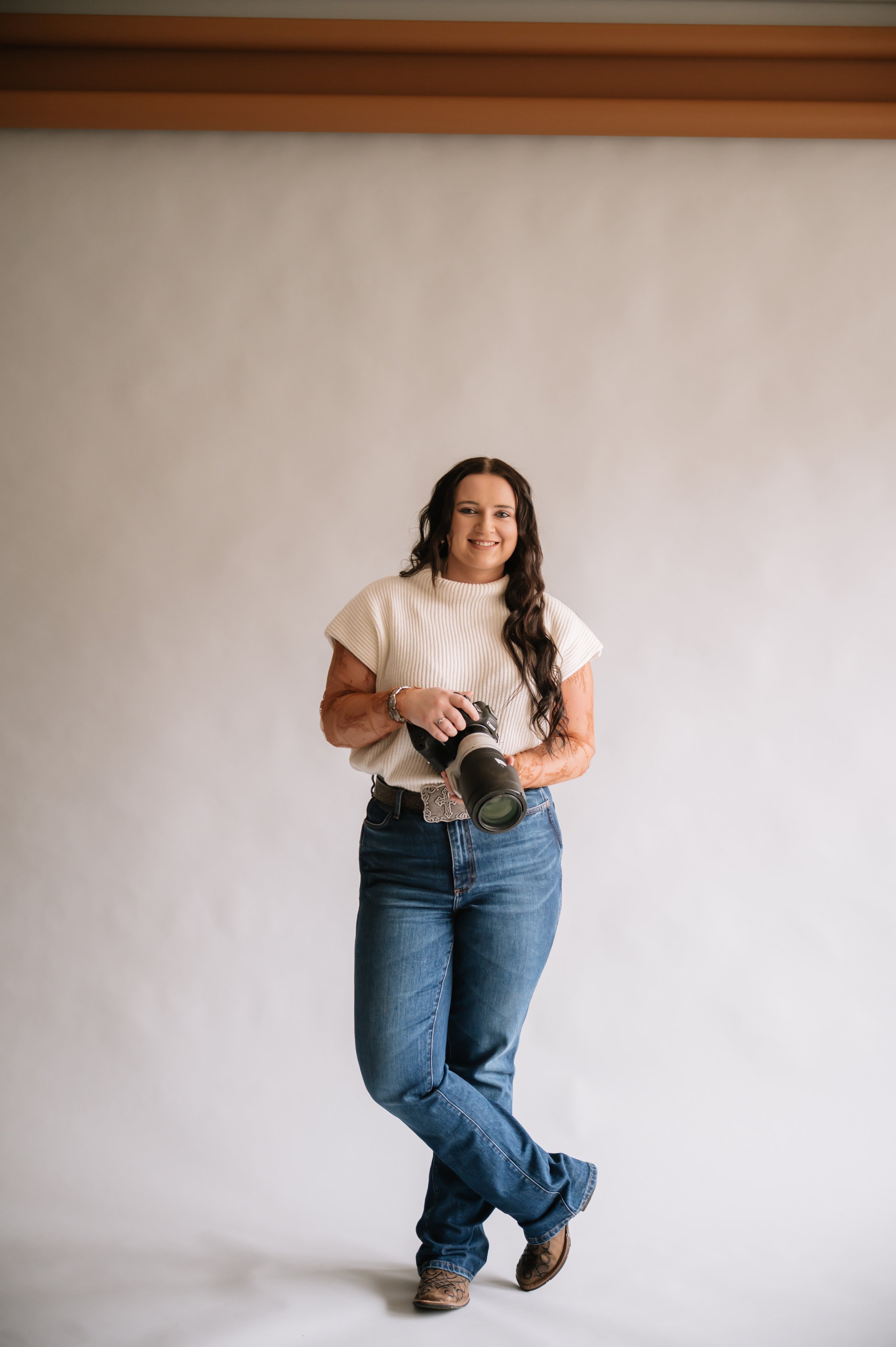 A woman with long dark hair, wearing a cream sweater, blue jeans, and cowboy boots, holding a camera, standing against a plain backdrop.