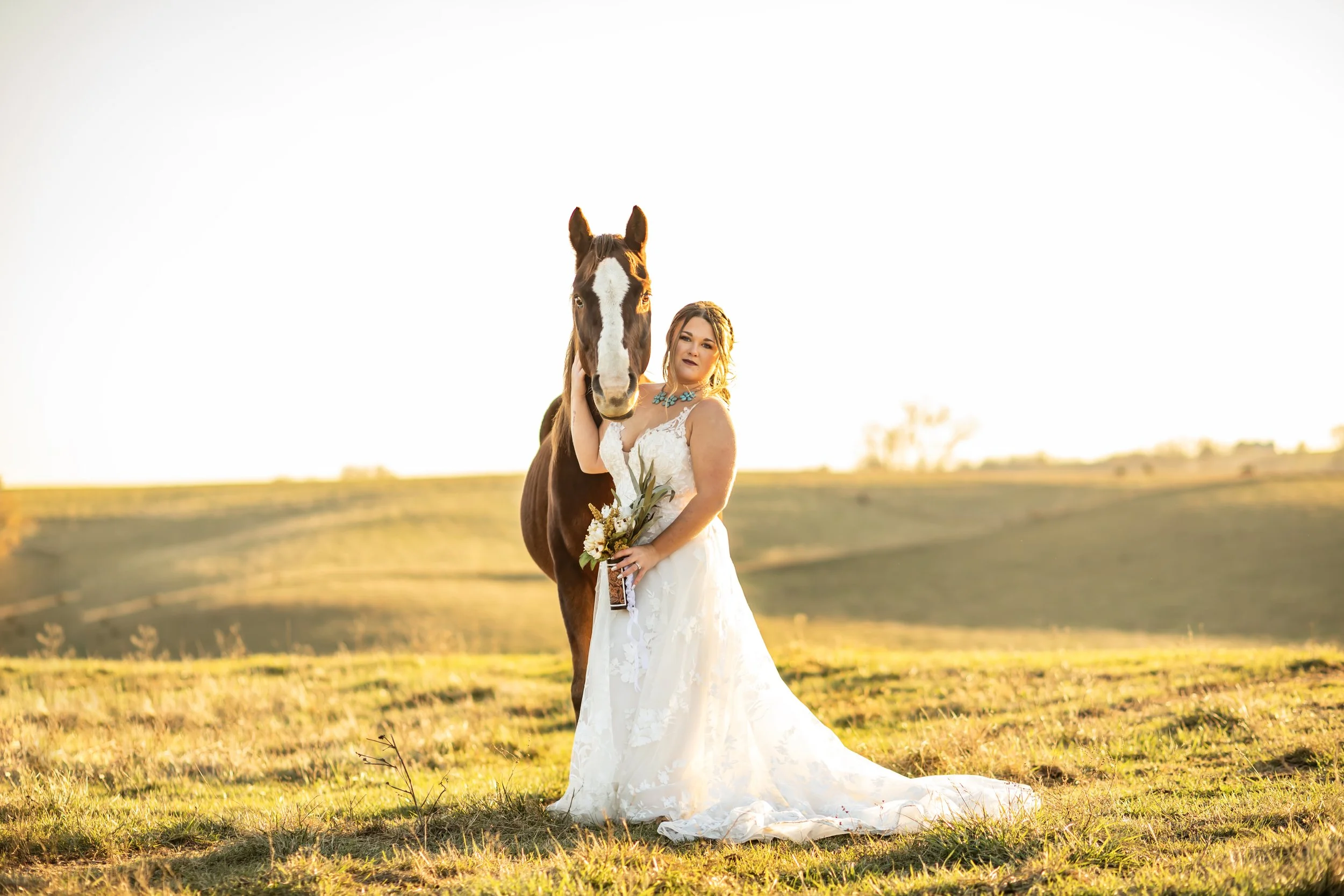 A woman in a wedding dress holding a bouquet standing next to a horse in an open field during sunset.