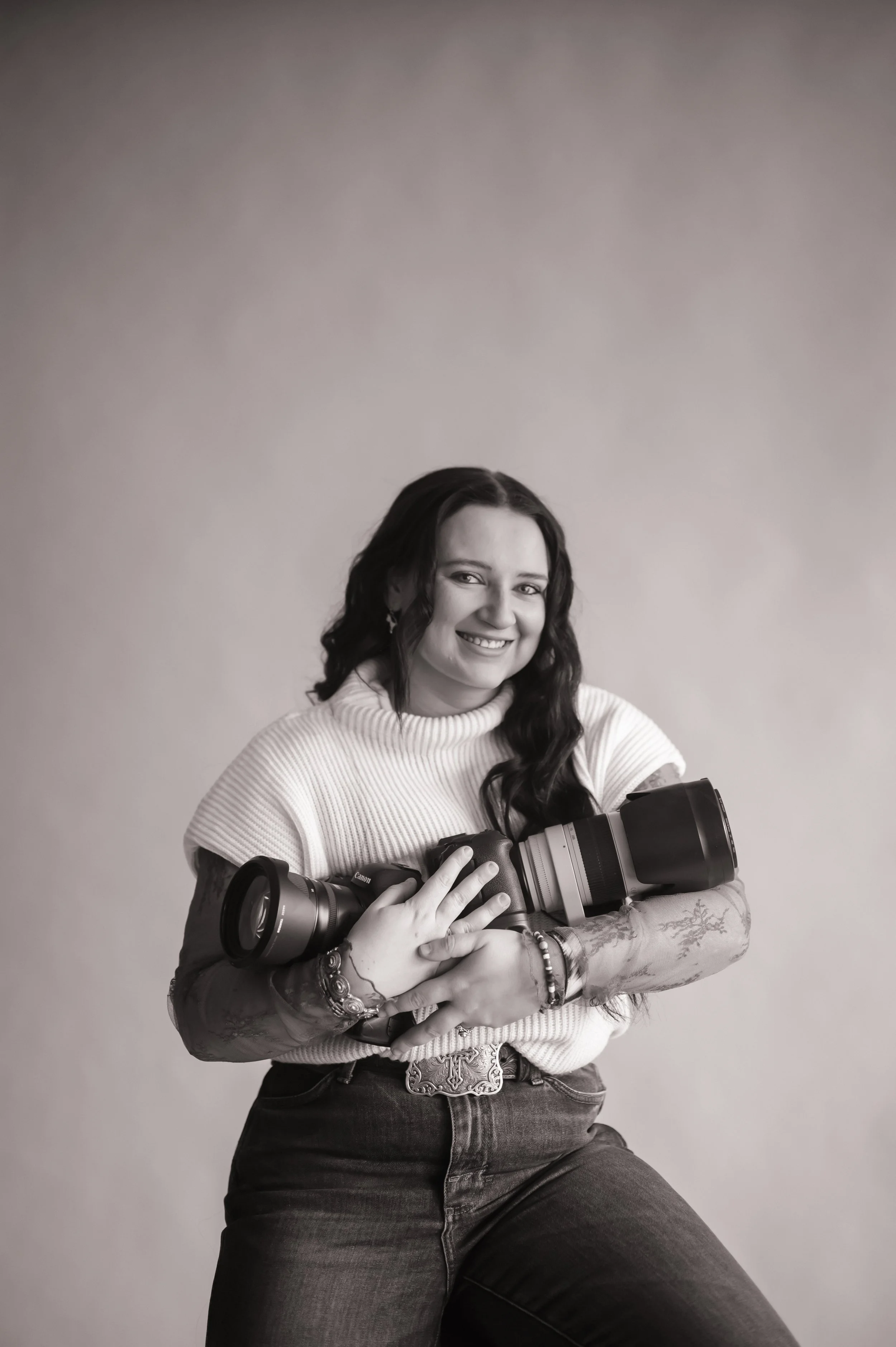 A woman with dark wavy hair smiling while holding a professional camera, wearing a white knitted sweater and jeans, against a plain backdrop.