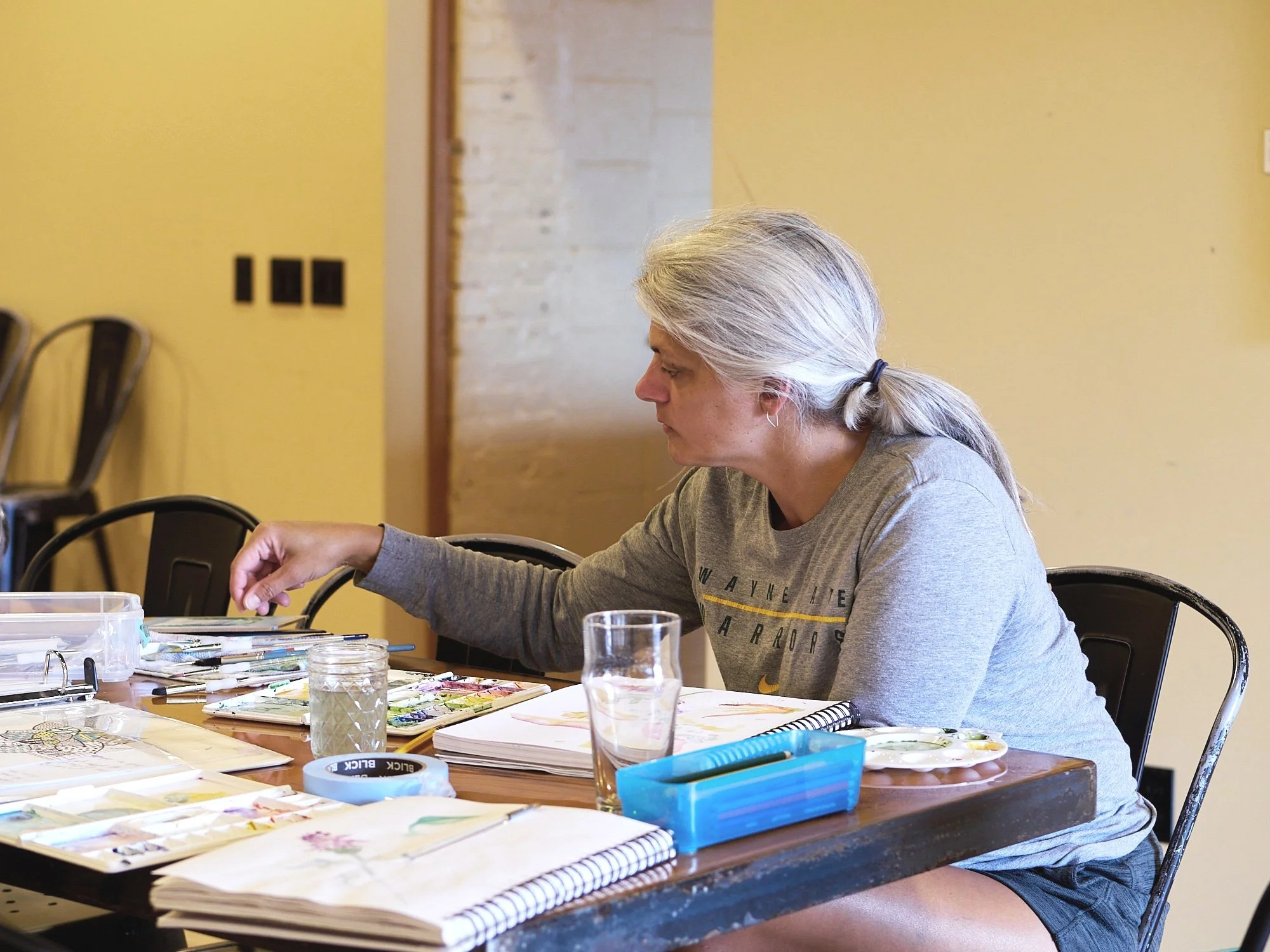 A woman with gray hair tied in a ponytail sitting at a table, working on watercolor painting supplies, surrounded by notebooks, watercolor palettes, jars, and brushes.