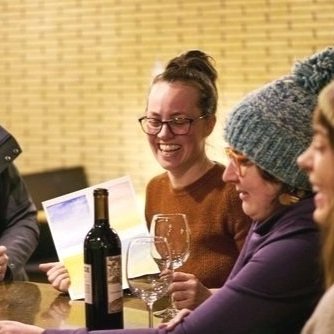 Four women sit at a table with a bottle of wine and glasses, smiling and engaging in conversation in a warmly lit indoor setting.