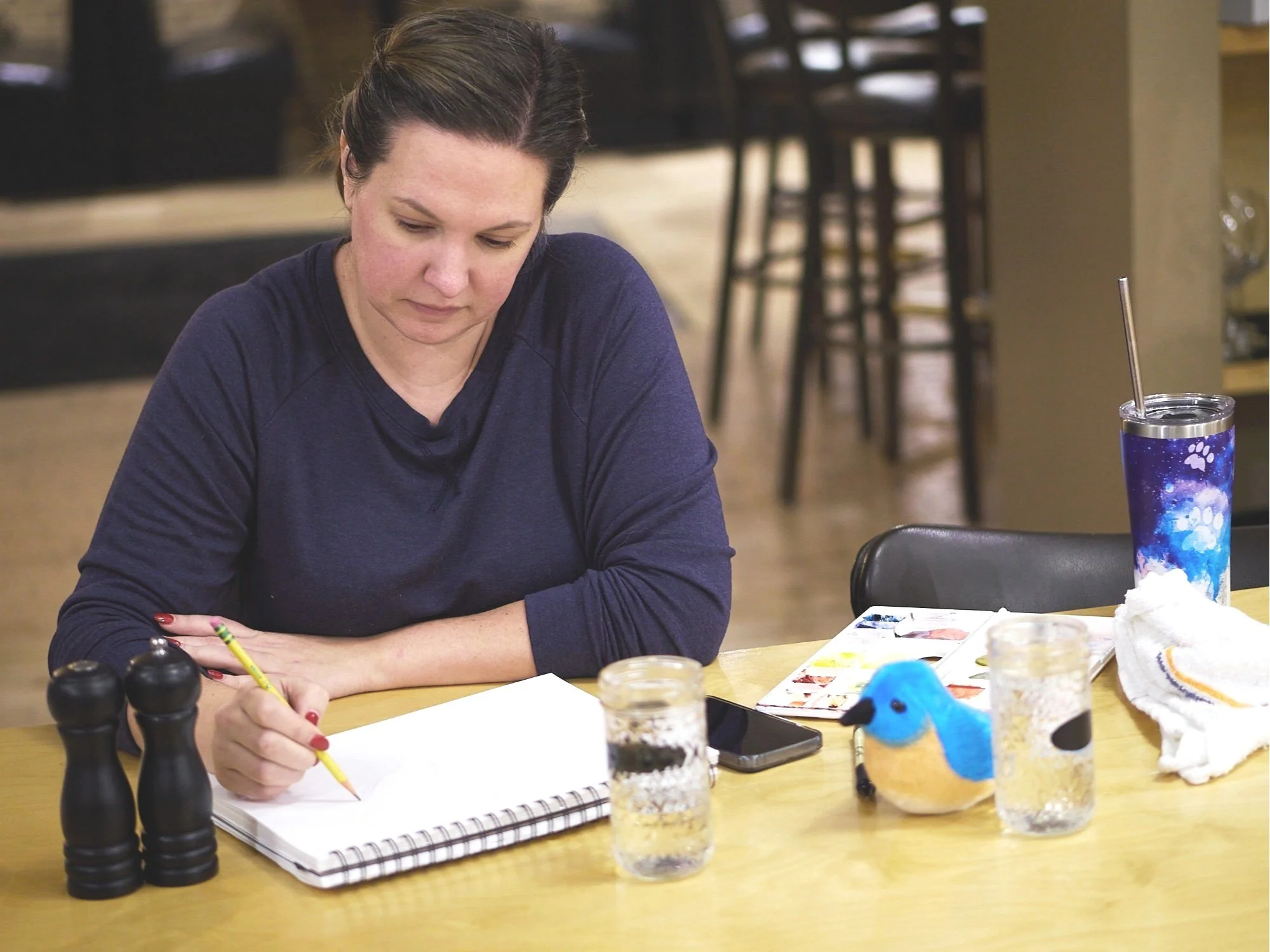 A woman with dark hair, wearing a navy blue shirt, sitting at a wooden table, drawing in a sketchbook with a yellow pencil. The table has various items, including a salt and pepper shaker, a plush blue and yellow bird, two glasses of water, a smartphone, a colorful tumbler, a white cloth, and a small picture book. The background shows a restaurant or café interior with chairs and tables.