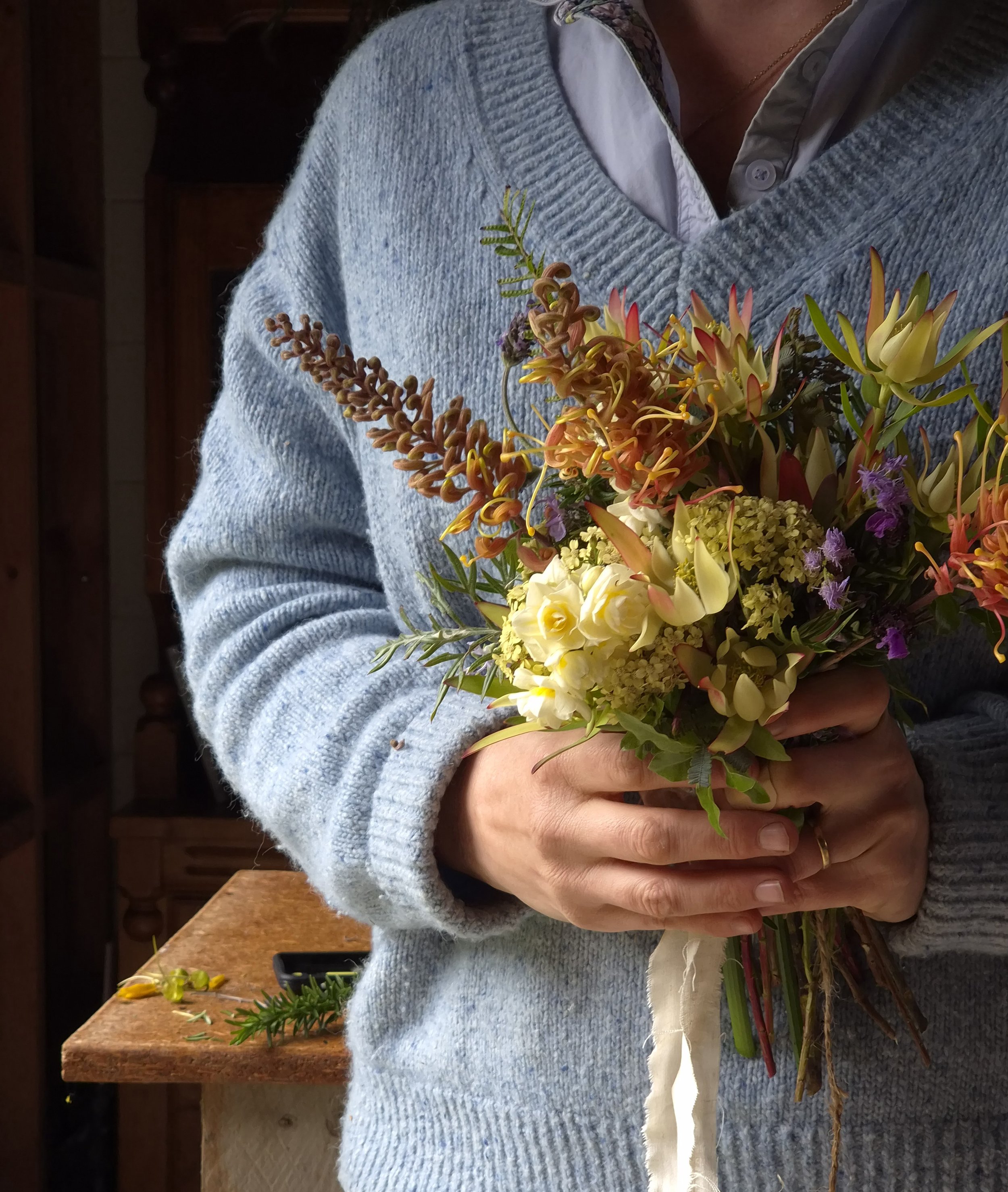 Person holding a bouquet of various colorful flowers in a room with a wooden shelf and table.