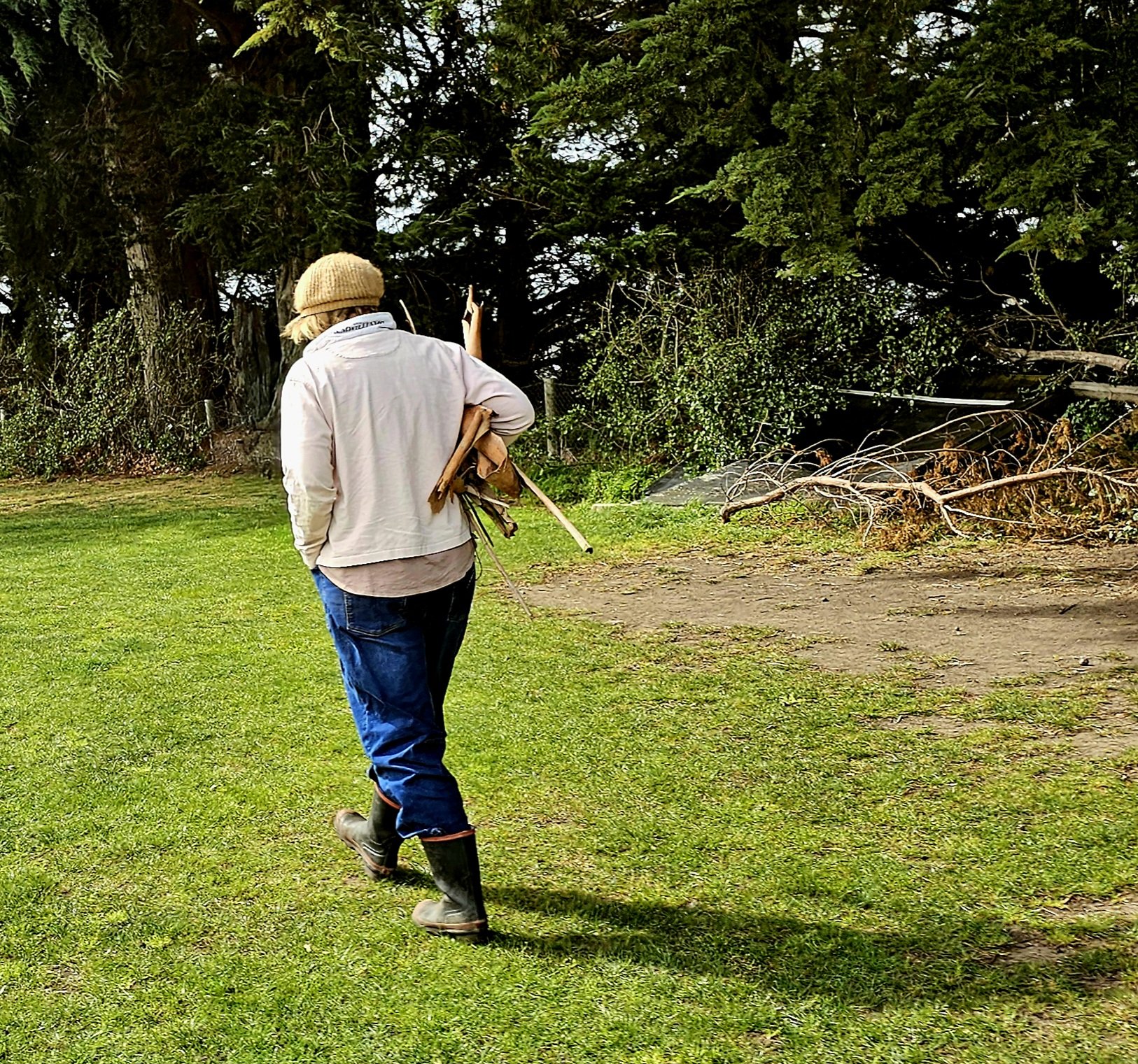 A person wearing a tan beanie, beige jacket, blue jeans, and black rain boots walking on a grassy area with a pile of branches and trees in the background.