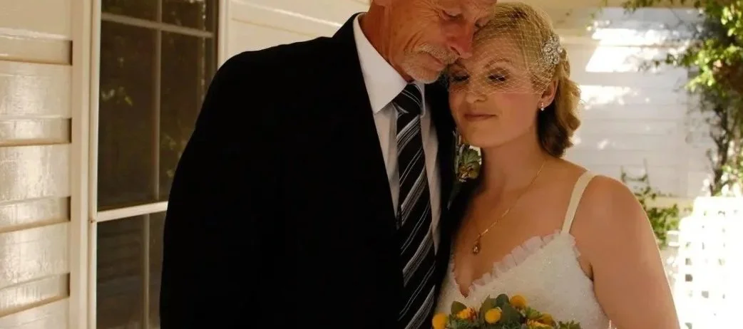 A bride and her father, standing close together at a wedding. The bride is holding a bouquet and wearing a lace wedding dress with a birdcage veil. The older man is in a black suit with a striped tie, and both are smiling softly.