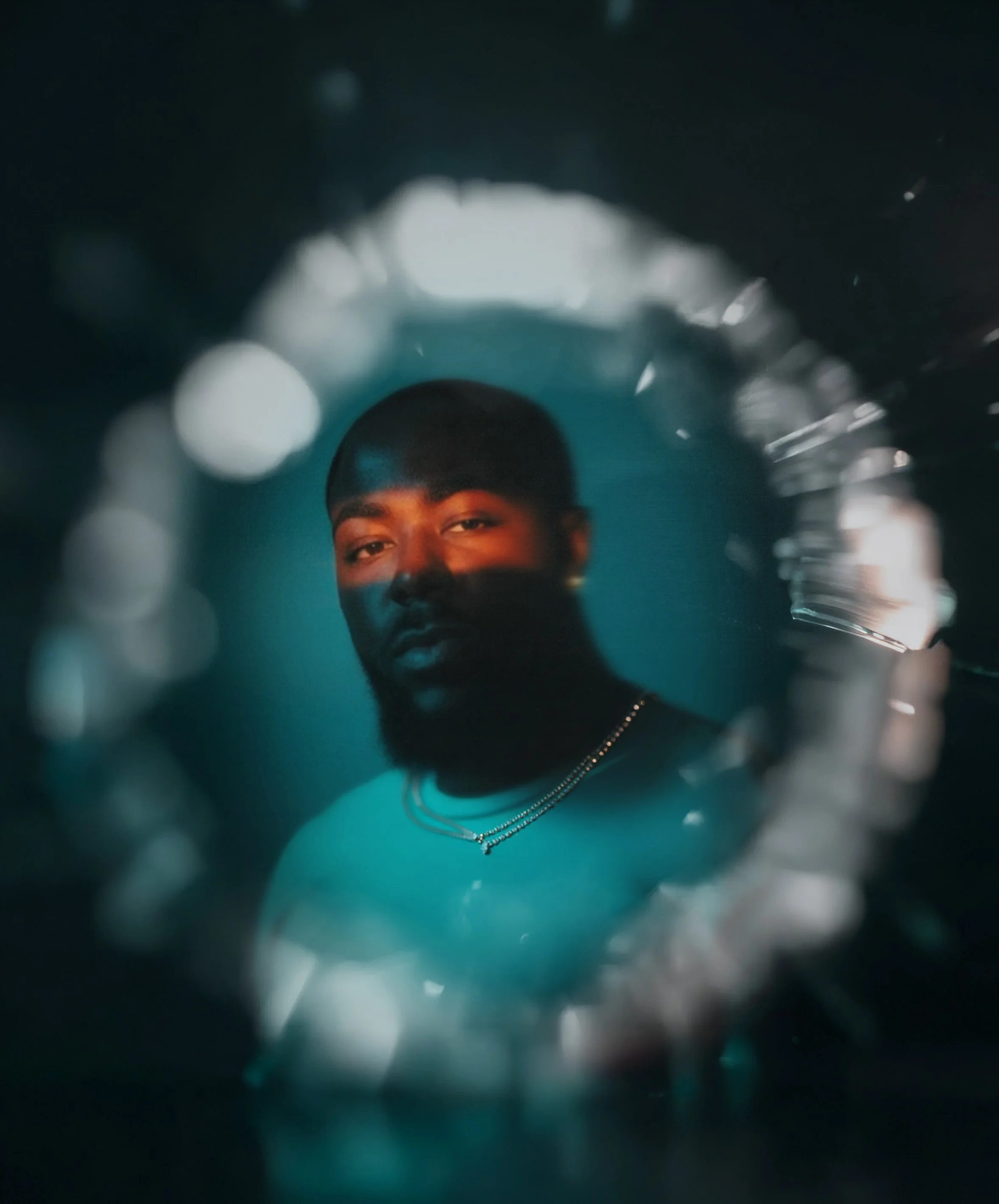 A portrait of a man with a beard and short hair, looking at the camera, visible through a circular reflective surface with a bokeh effect around it.