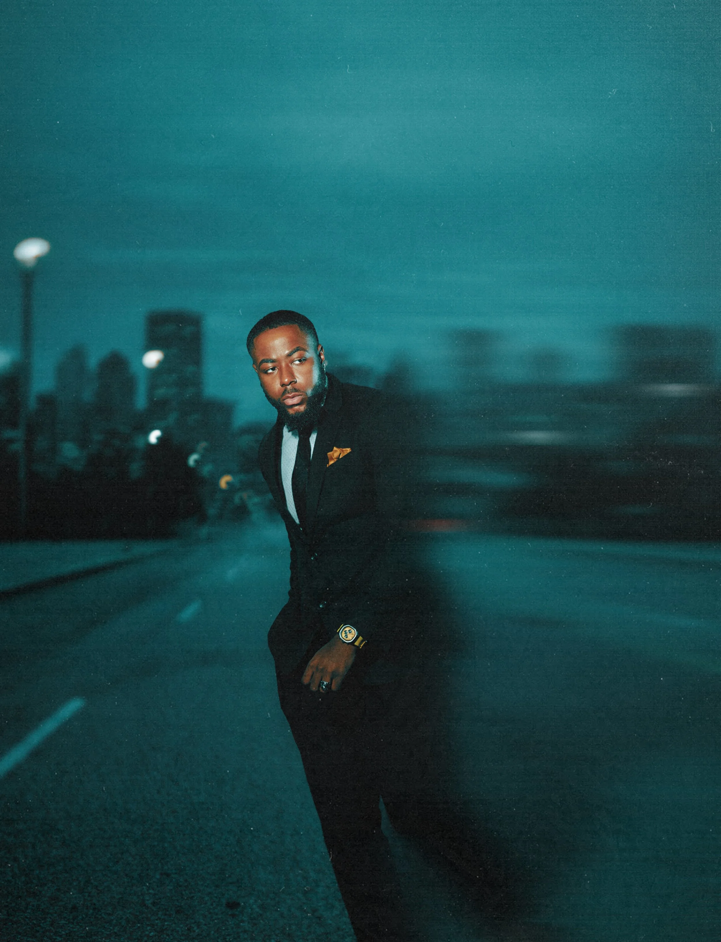 A man wearing a black suit, white shirt, and black tie standing on an empty city street at night with blurred cityscape in the background.