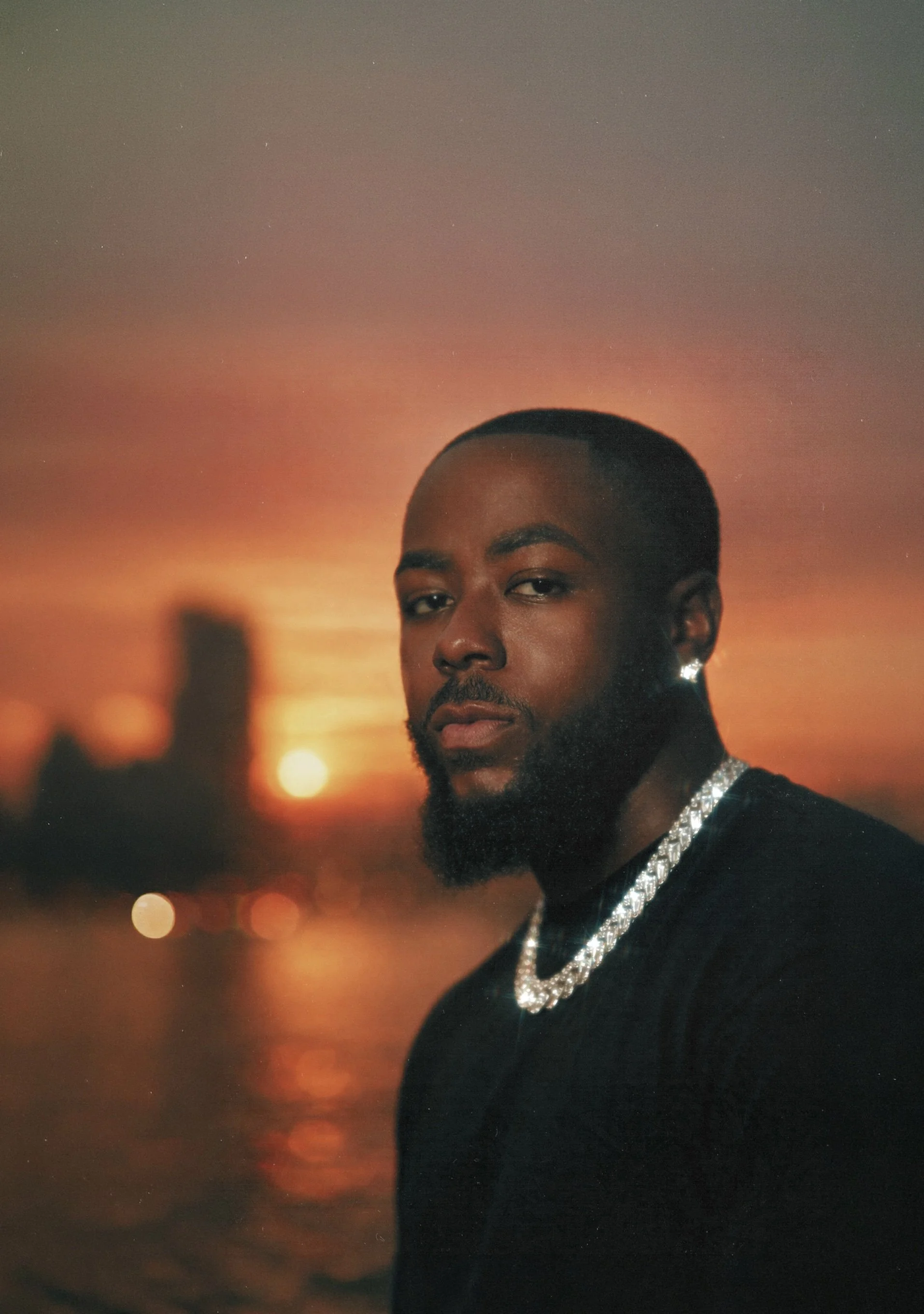 A man with a beard and short hair posing outdoors at sunset, wearing a black shirt and a diamond chain necklace, with a city skyline blurred in the background.