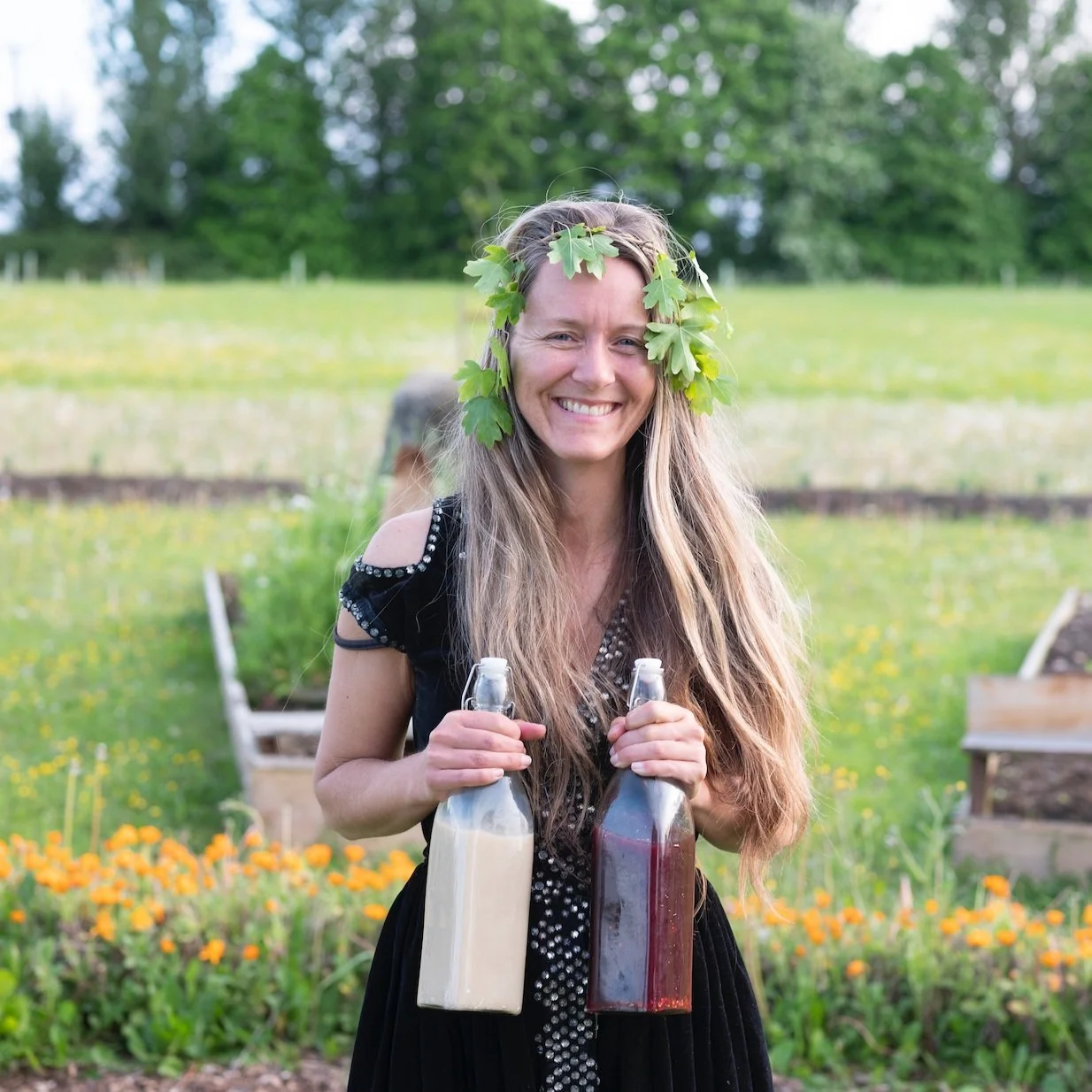 Woman with long hair and leaf headband holding two bottles, one with white liquid and the other with dark liquid, standing in a garden with trees and flowers in the background.