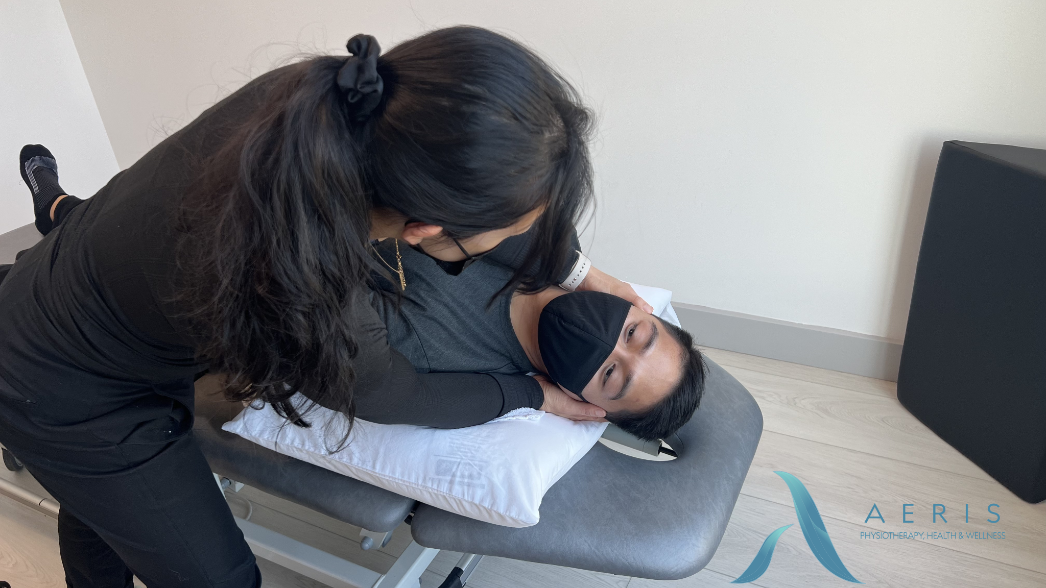 A woman with long dark hair and glasses performs a physical therapy exercise on a man lying on a therapy table. The man is wearing a black mask and a gray shirt, and the woman is dressed in black. The setting appears to be a physiotherapy clinic with a pillow under the man's head and a logo that reads "AERIS Physiotherapy, Health & Wellness" in the bottom corner.