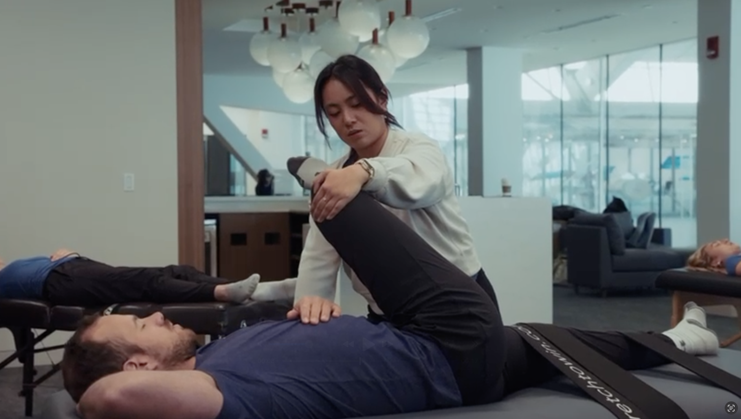 A woman performs a physical therapy exercise on a man lying on a treatment table in a modern, well-lit rehabilitation facility.