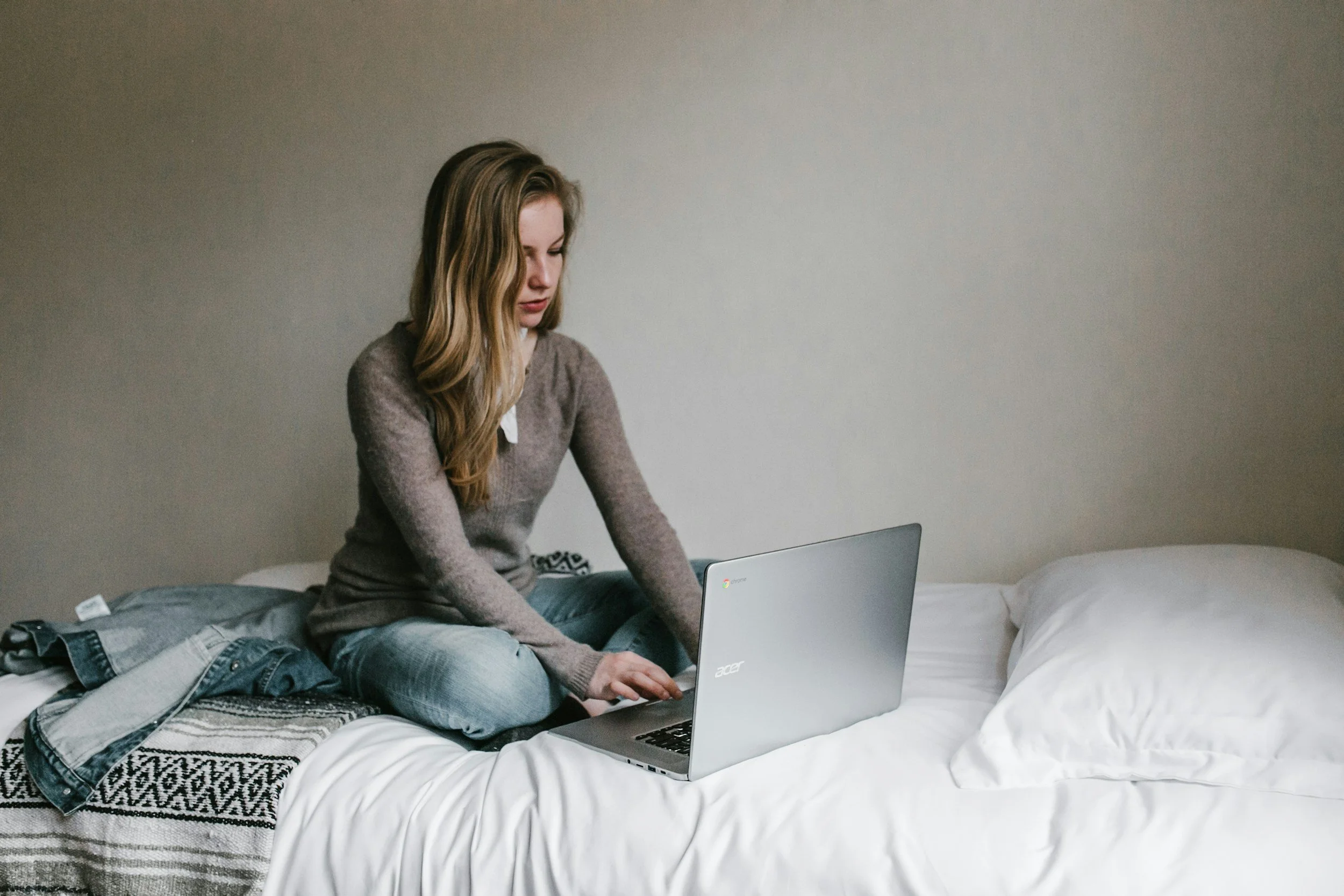 A young woman with long blonde hair is sitting on a bed with a white sheet and pillow, working on a silver laptop. She is wearing a brown sweater and light blue jeans, and there are some clothes, including a denim jacket, on the bed beside her. The background is a plain gray wall.