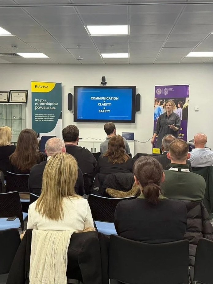 A woman is giving a presentation to an audience seated in rows. The screen behind her displays the words 'Communication Clarity Safety.' There are banners on either side of the screen, one for Aviva and another with a university logo.