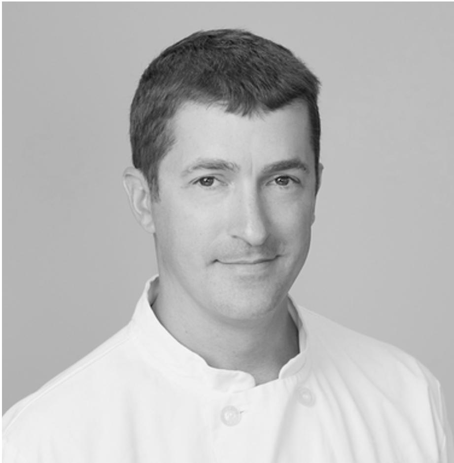 A male chef in a restaurant, wearing a white chef's coat, apron, and cap, sitting at a table with a glass of beer in front of him, inside a rustic wooden interior.