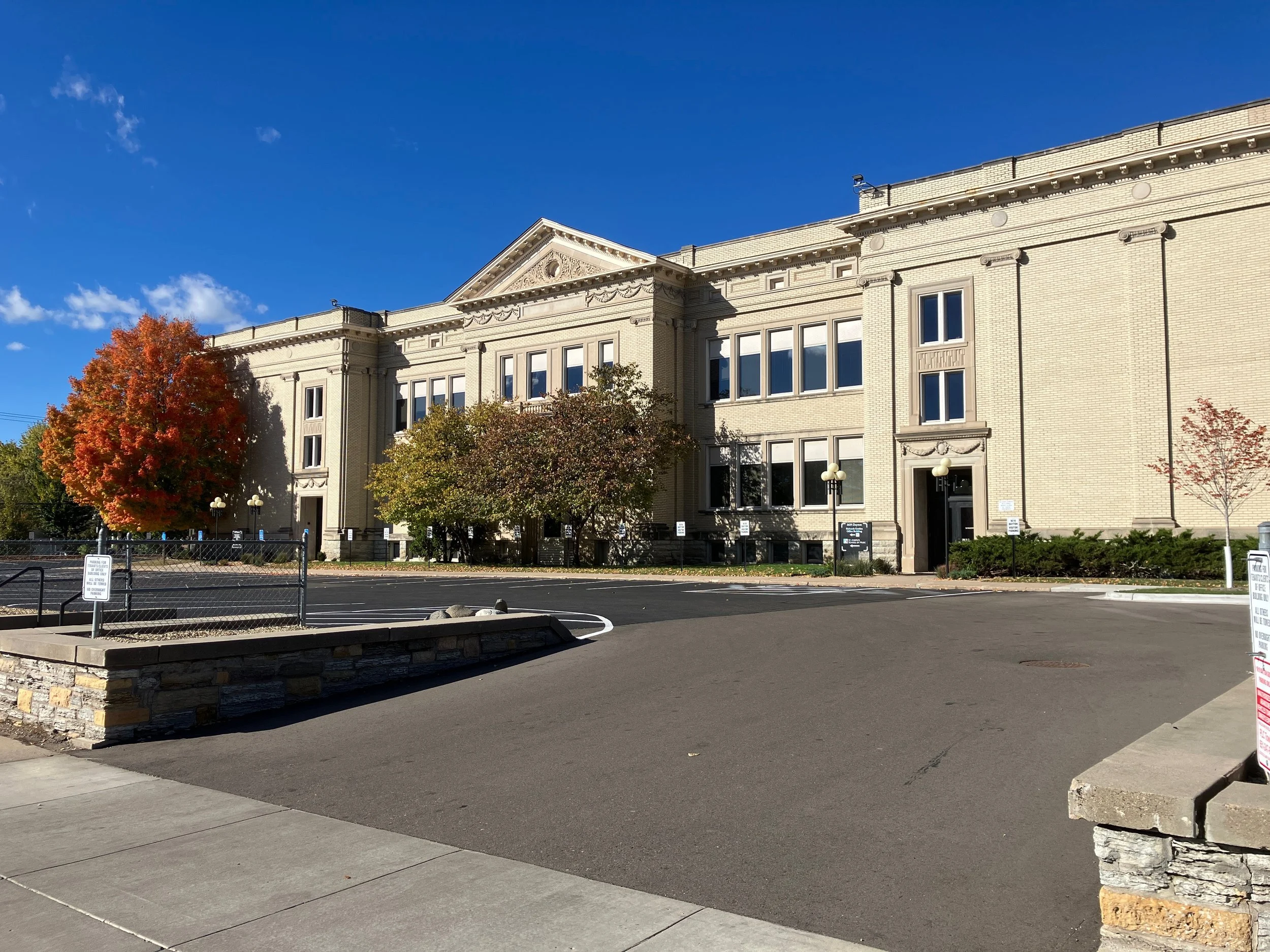 A large, historic-style brick building with multiple windows and decorative architectural details, surrounded by autumn trees and a parking lot under a clear blue sky.