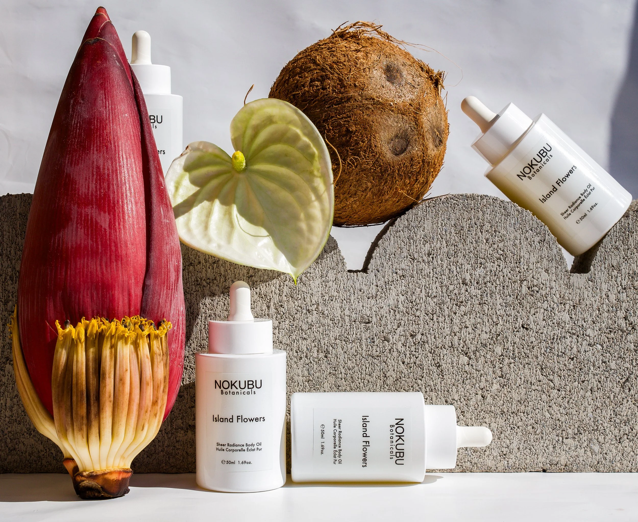 A close-up of a sliced tropical flower, a large root, and skincare bottles on a textured gray surface. The flower has peach and yellow petals with stalks and center. The skincare bottles are white with black text, labeled 'NOKUBU Botanicals Island Flowers Sheer Radiance Body Oil' and are arranged among the flowers and roots.