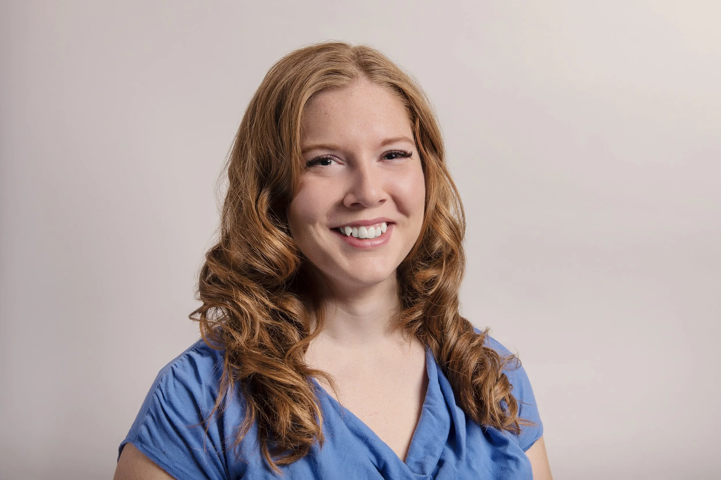 A young woman with curly red hair smiling, wearing a blue top, against a plain light background.