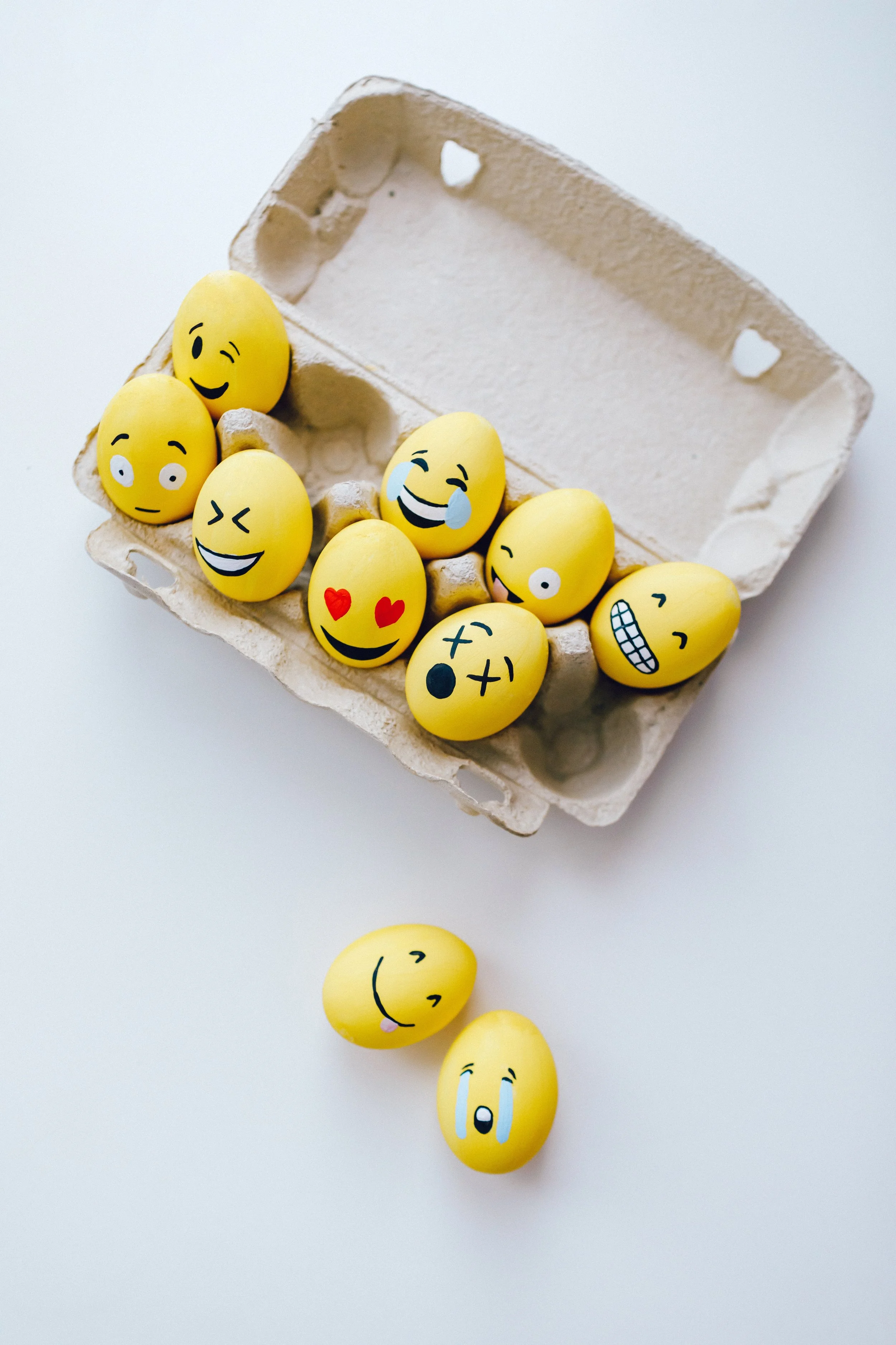 Yellow eggs decorated with various cartoon smiley face stickers, some with tears, hearts, or winking expressions, in an egg carton and on a white surface.